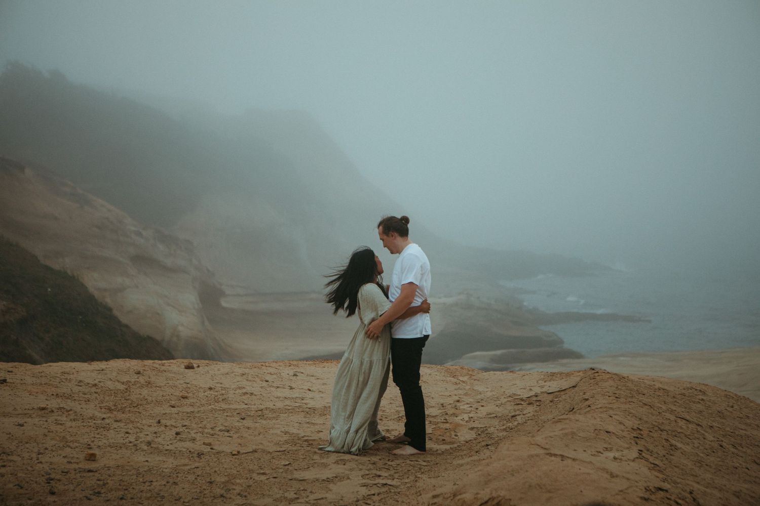 Couple embracing romantically on a foggy sandy beach with misty coastal cliffs in the background.