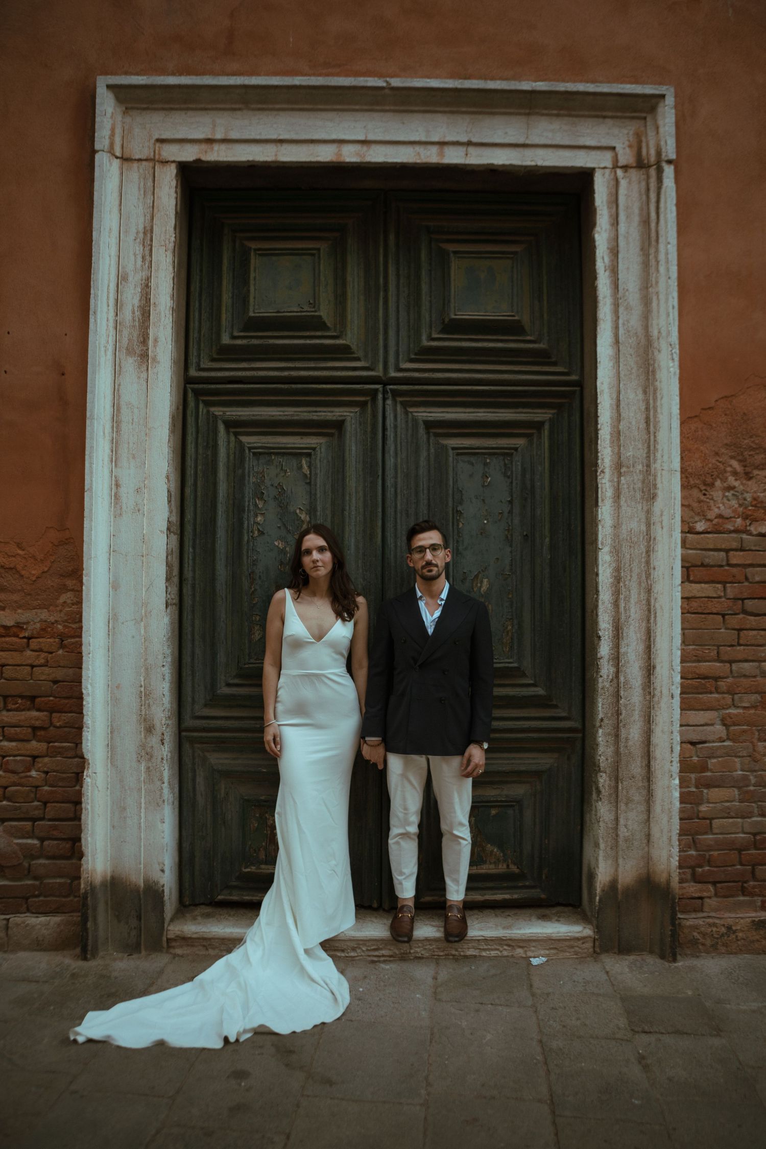 Bride in white gown and groom in dark blazer pose before ornate green door in Venice.