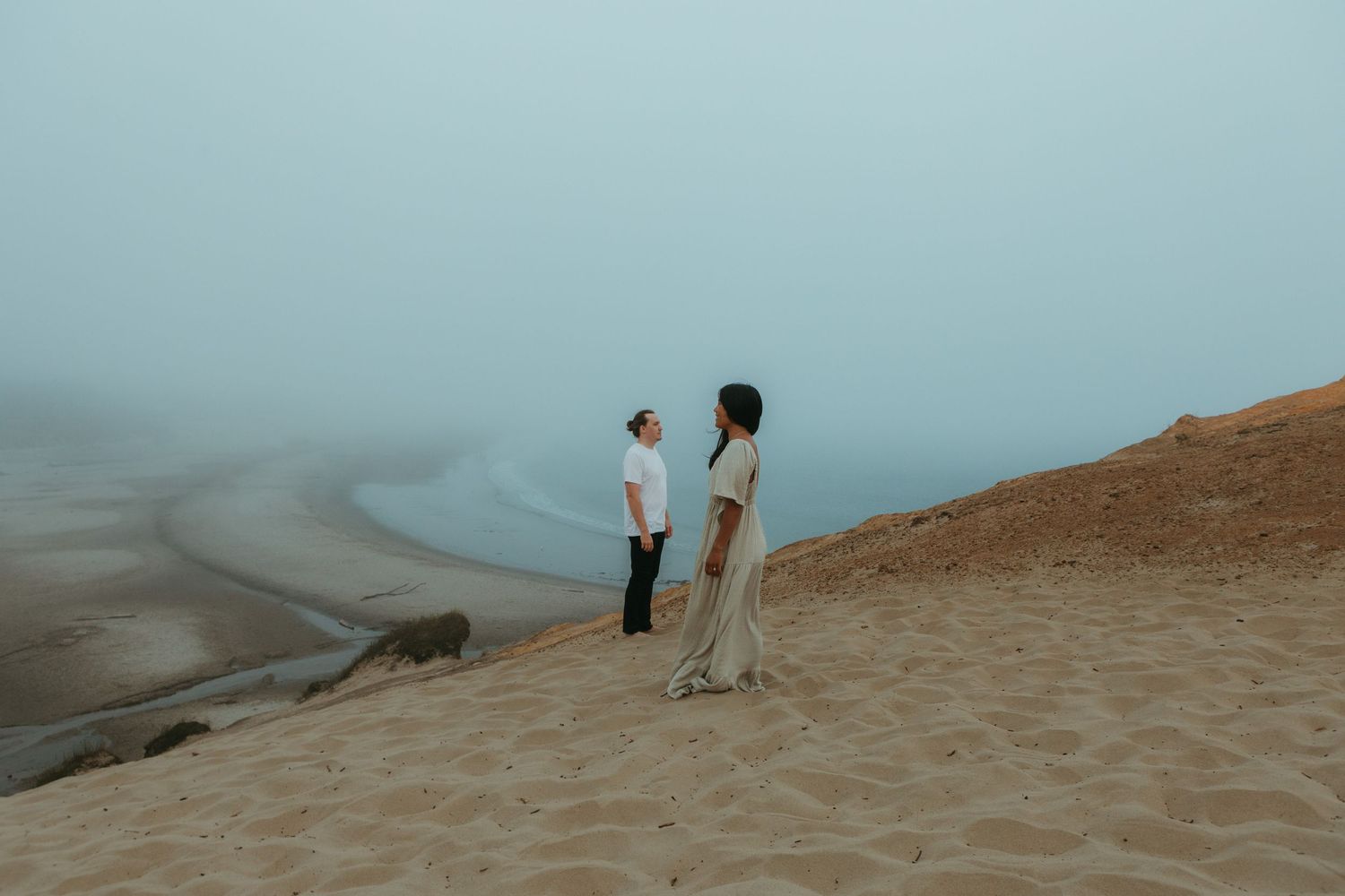 Two people stand on a sandy dune overlooking a misty coastal landscape on a foggy day.