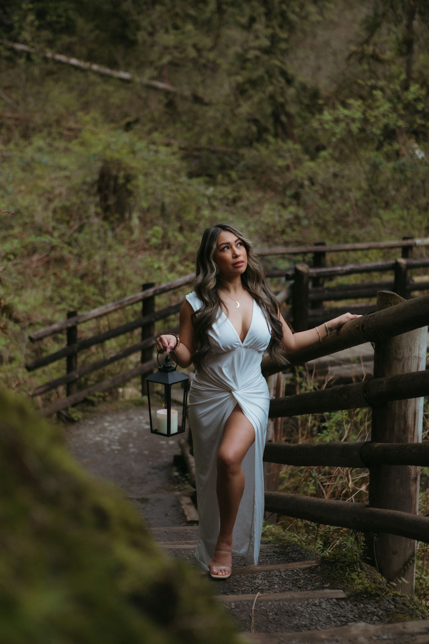 A figure in a flowing white dress poses elegantly along a rustic wooden fence pathway in a lush forest setting.
