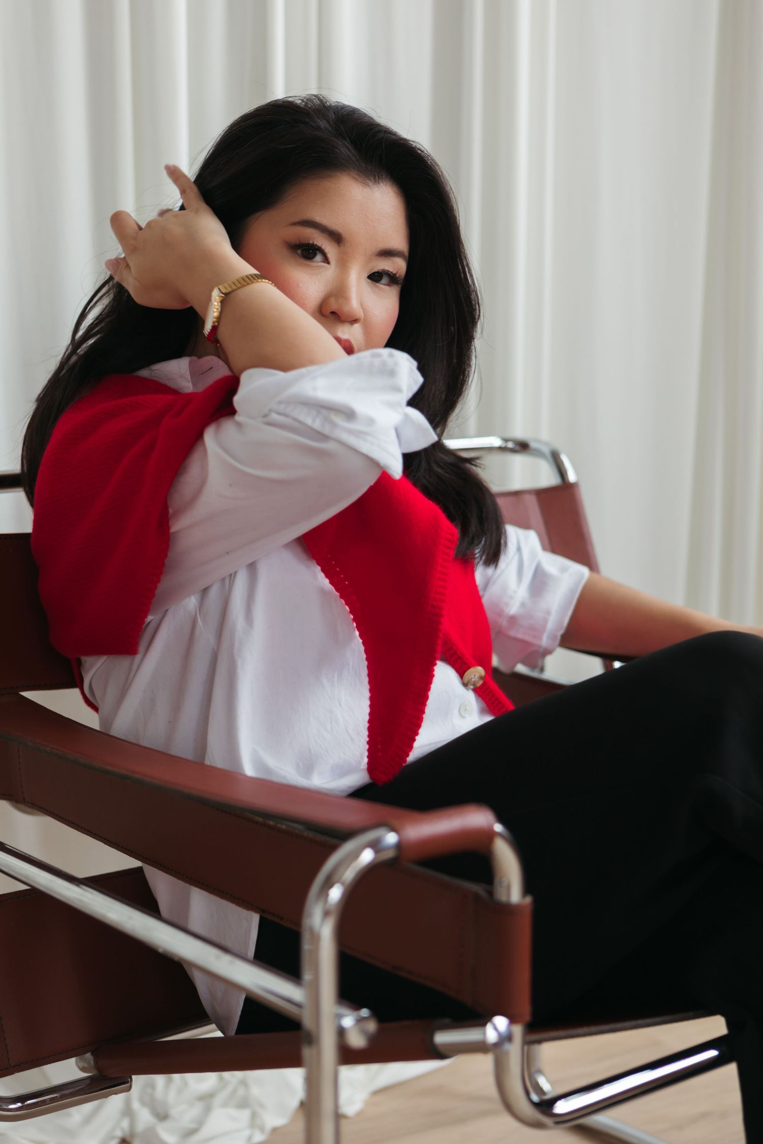 Woman with long dark hair in a white shirt and red sweater draped over her shoulders, posing in a chrome and leather chair.