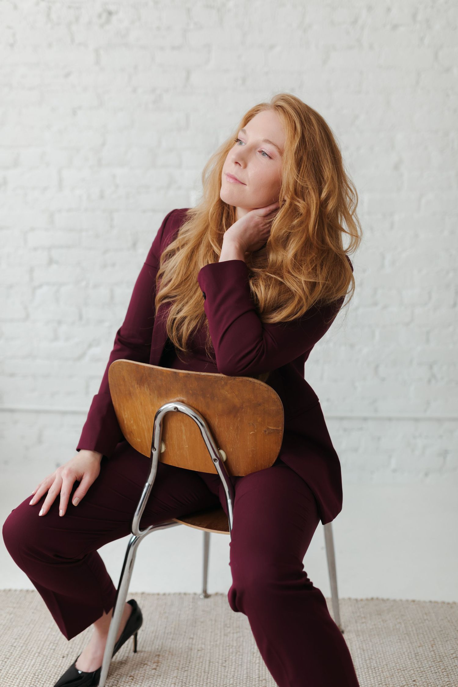 Person in burgundy dress lounges expressively on modern chair against white brick wall in artistic portrait series.