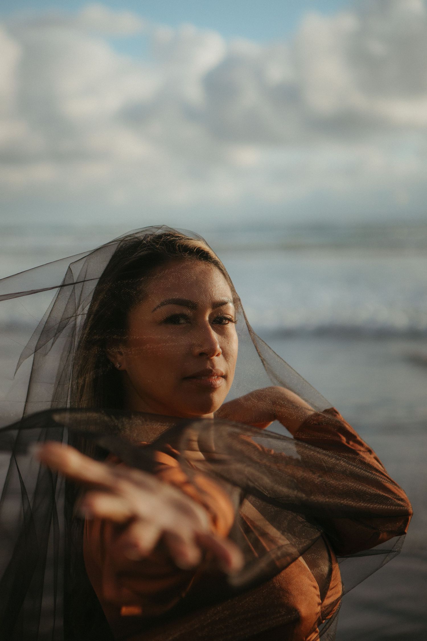 Atmospheric beach portrait with sheer fabric flowing in the wind against a cloudy ocean backdrop.
