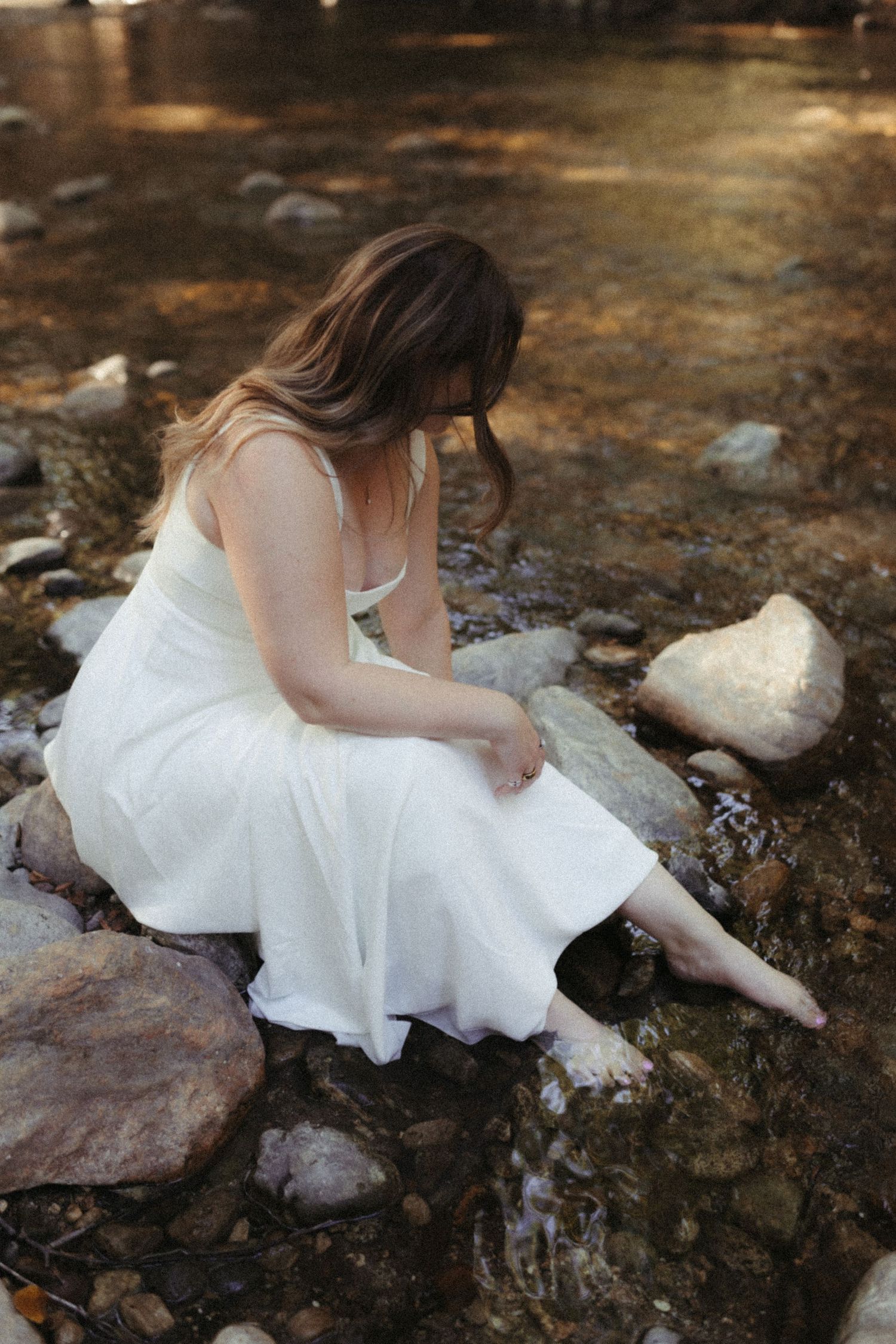 A flowing white dress brushes against mossy rocks near a stream in an ethereal woodland setting.