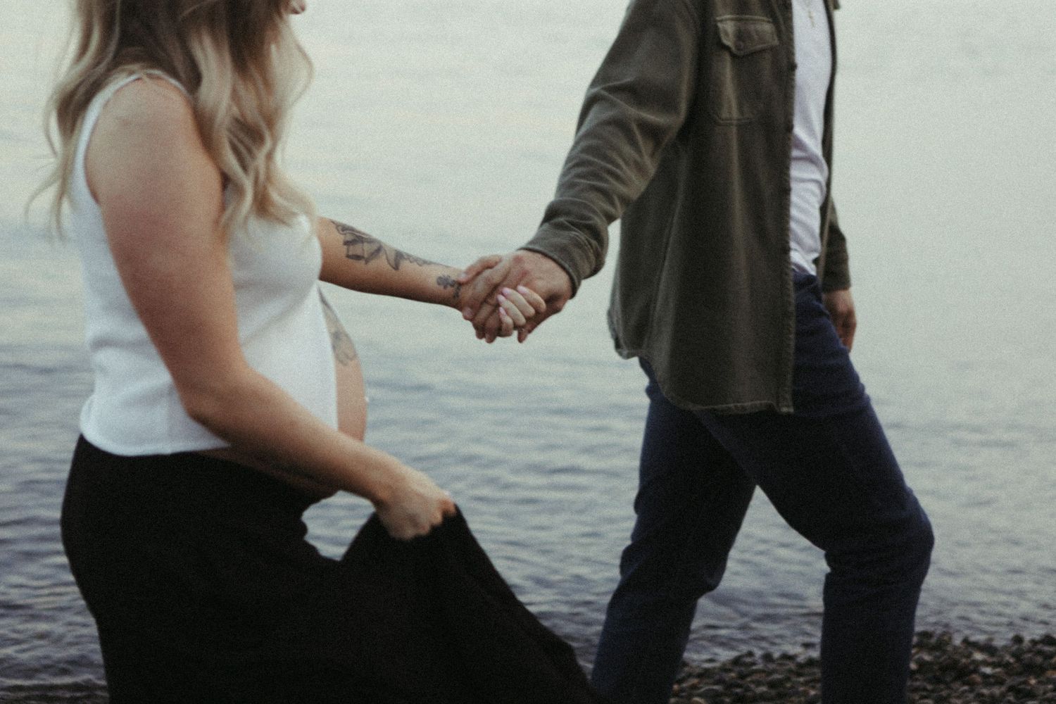 Couple in formal attire holds hands and dances together on a beach at sunset with ocean waves in the background.