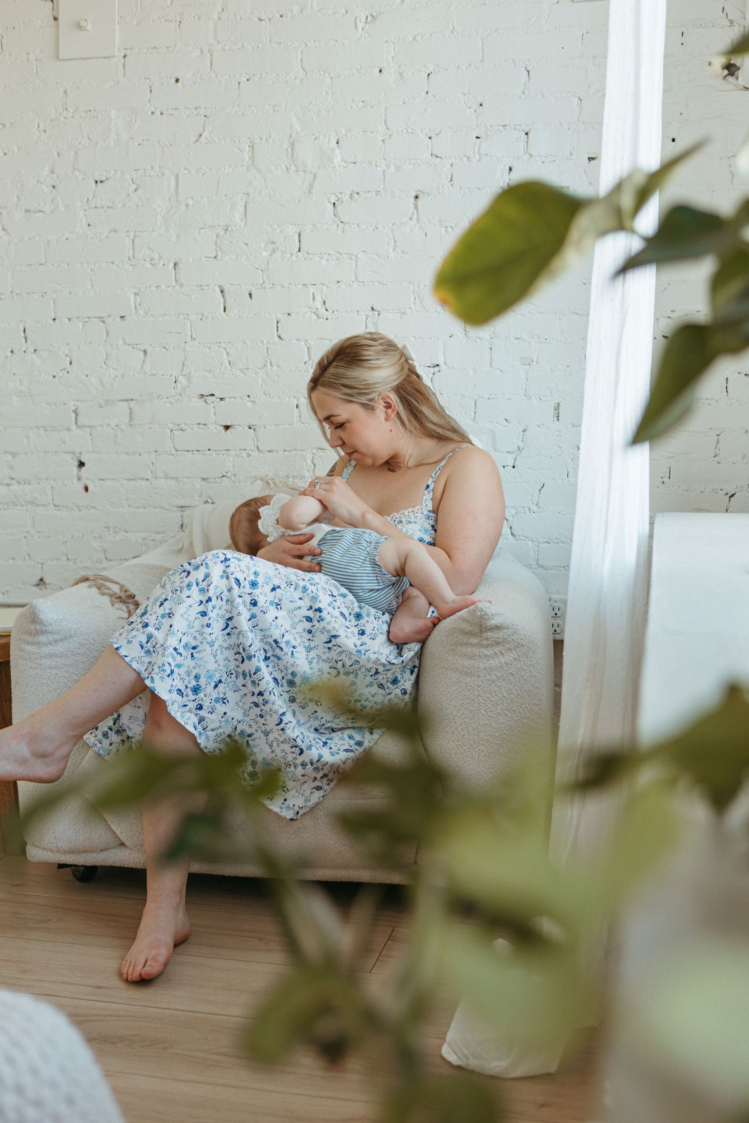 A mother breastfeeding her newborn baby while sitting on a couch in a bright white room.