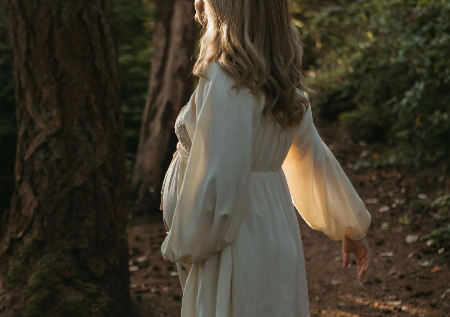A person in a flowing cream-colored dress walks through a sunlit wooded forest path.