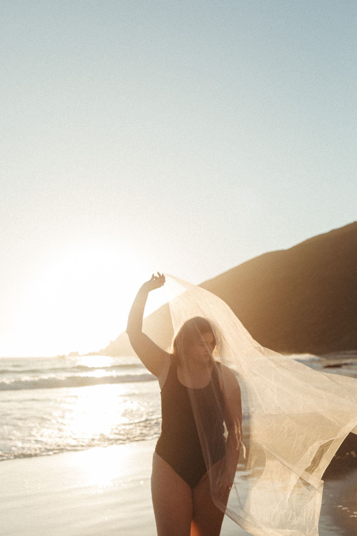 A flowing white fabric catches the sunlight on a beach during golden hour, creating an ethereal seaside scene.