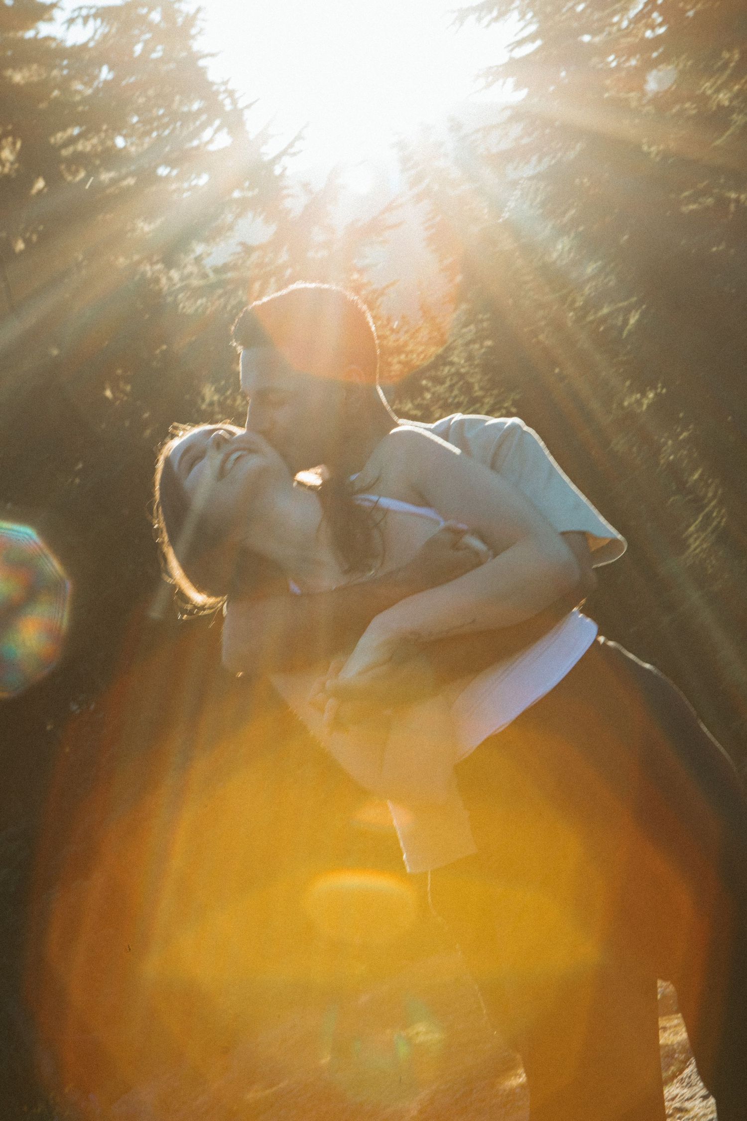 Couple embraces in a sunlit forest with dramatic lens flare creating a warm golden glow.