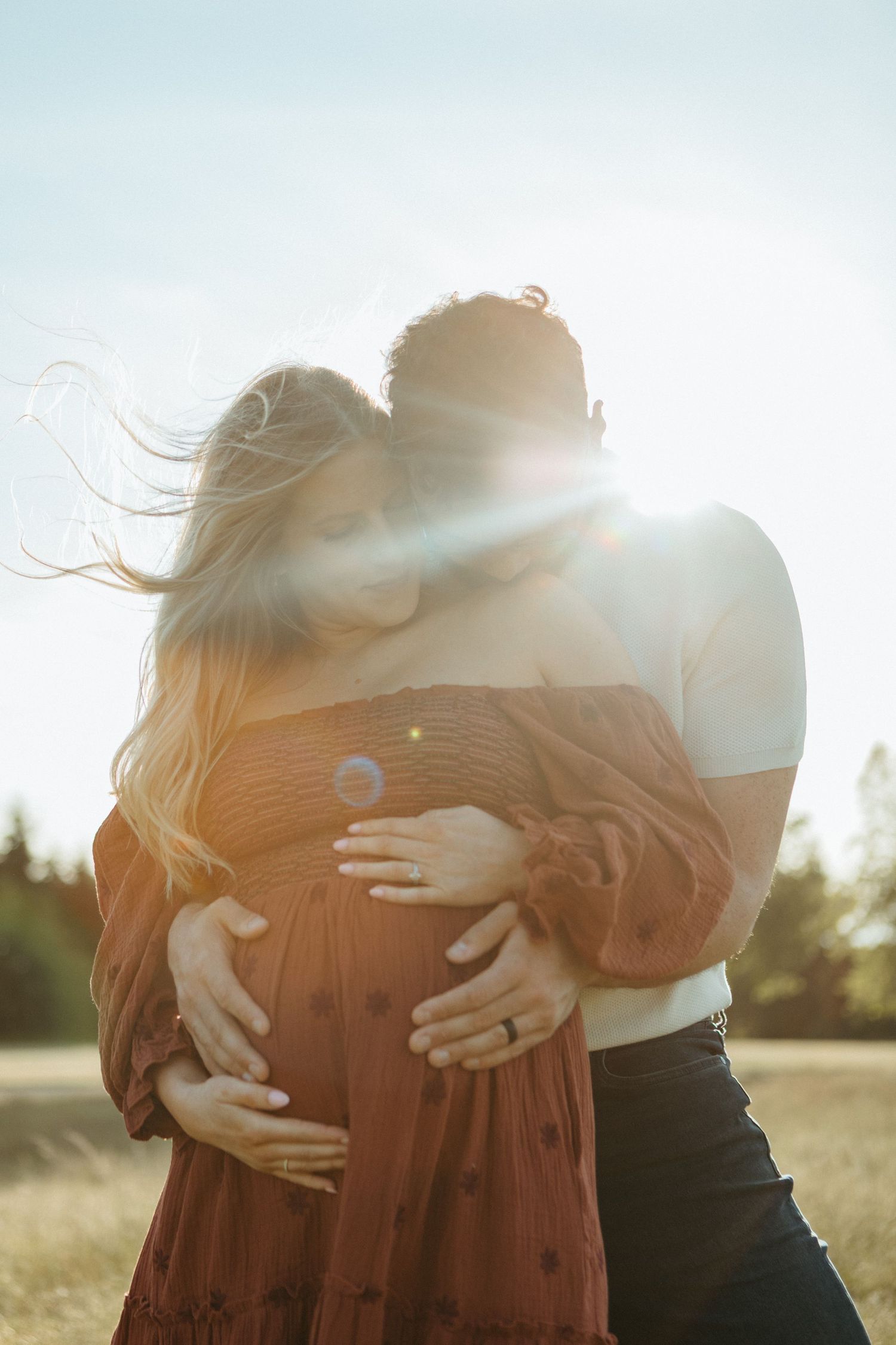 A tender embrace captured in golden sunlight with lens flare creates a dreamy romantic moment in a field at sunset.
