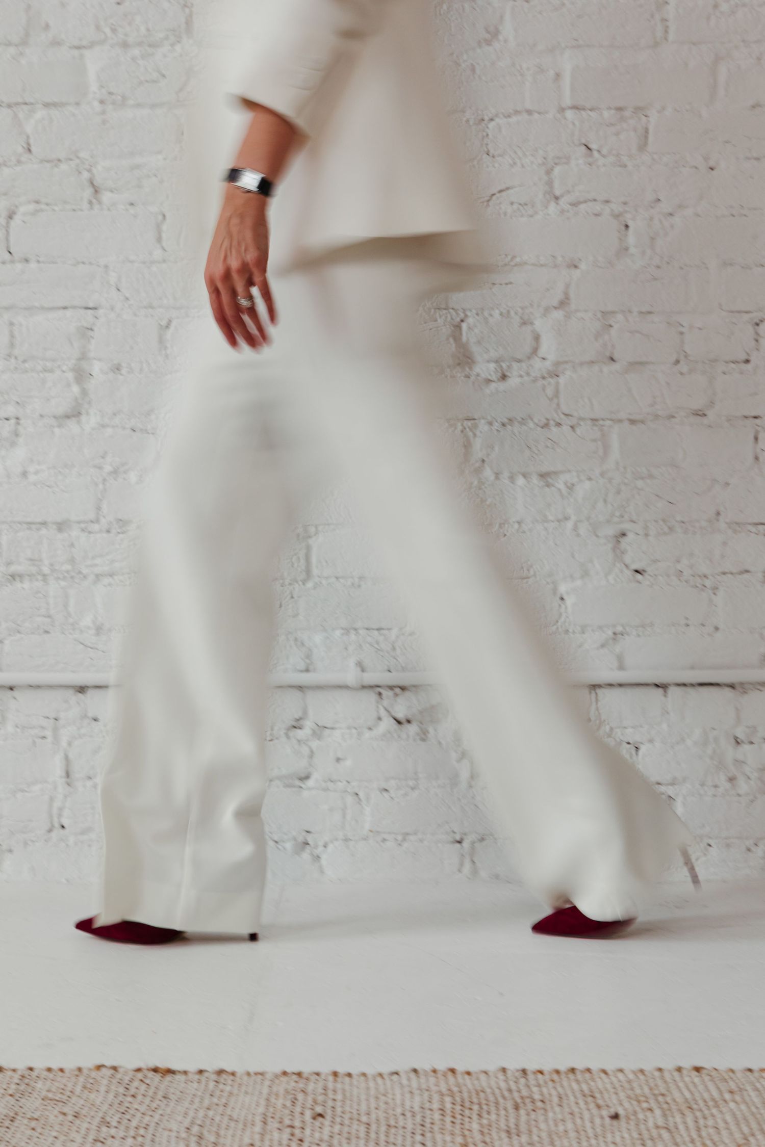 Cropped view of legs wearing white wide-leg pants and burgundy shoes walking against a white brick wall background.