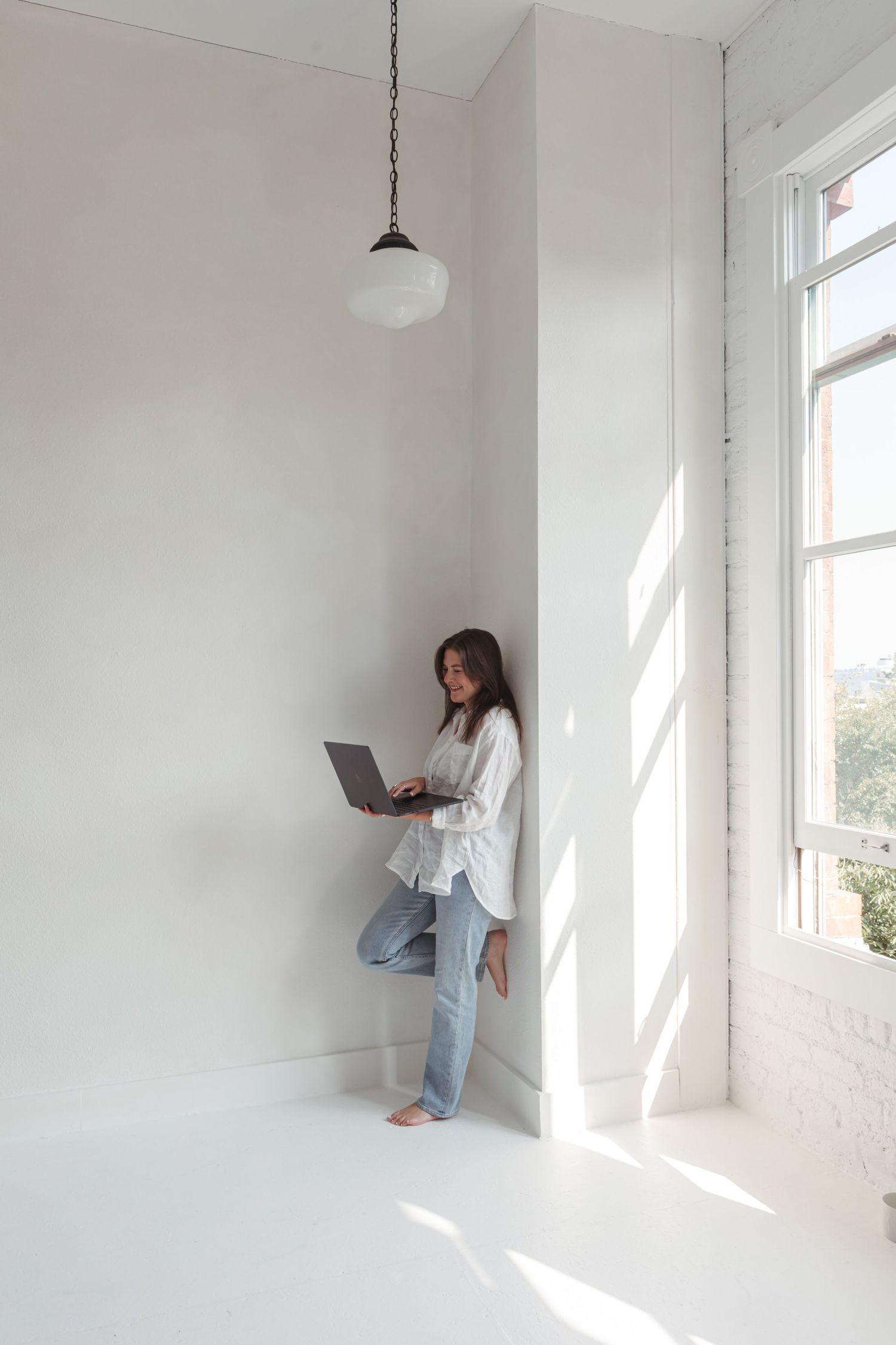 Person in white shirt and jeans sits by bright window in minimalist white room working on laptop computer.