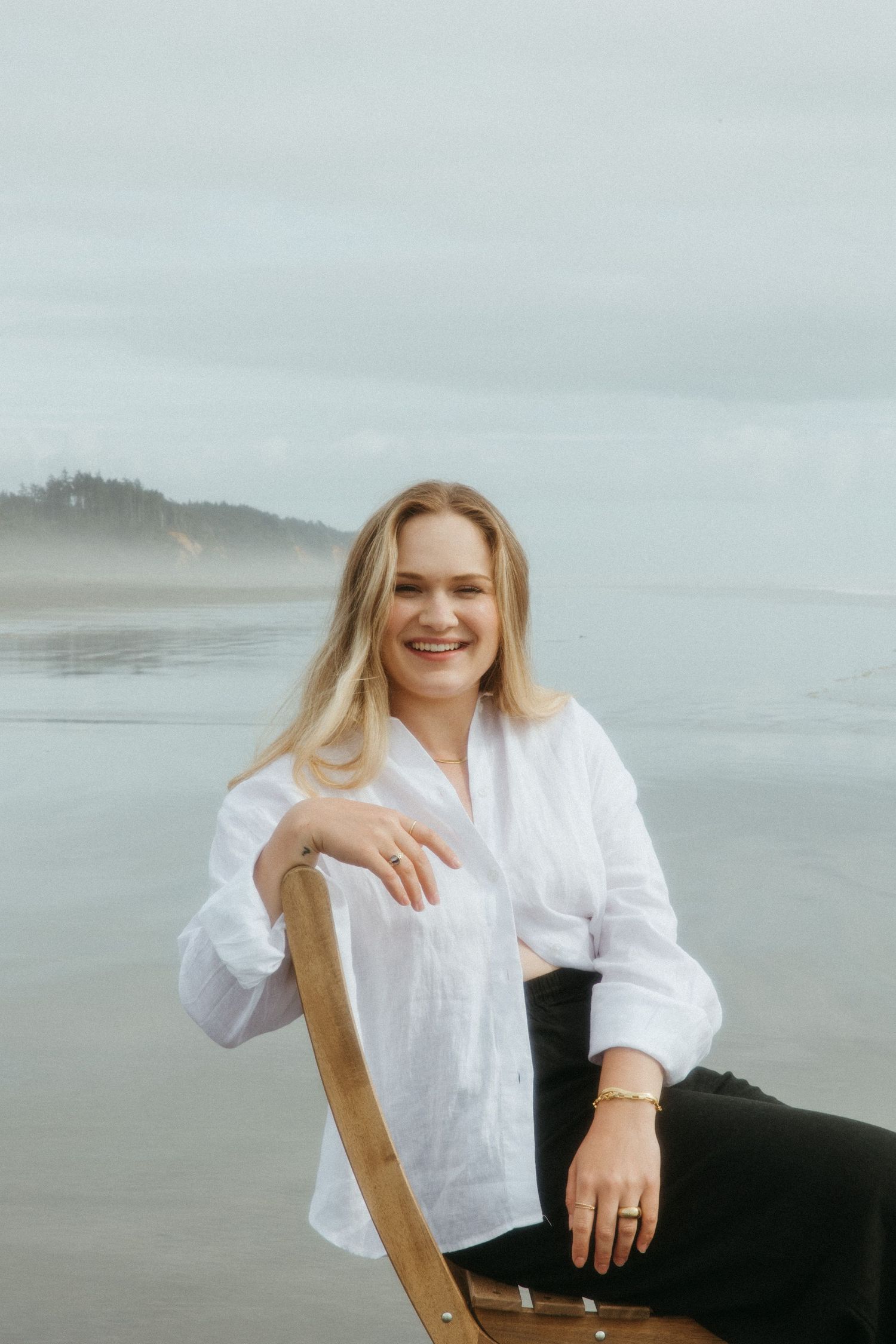 Smiling blonde woman in white linen shirt sits on wooden chair on misty beach with calm ocean backdrop.