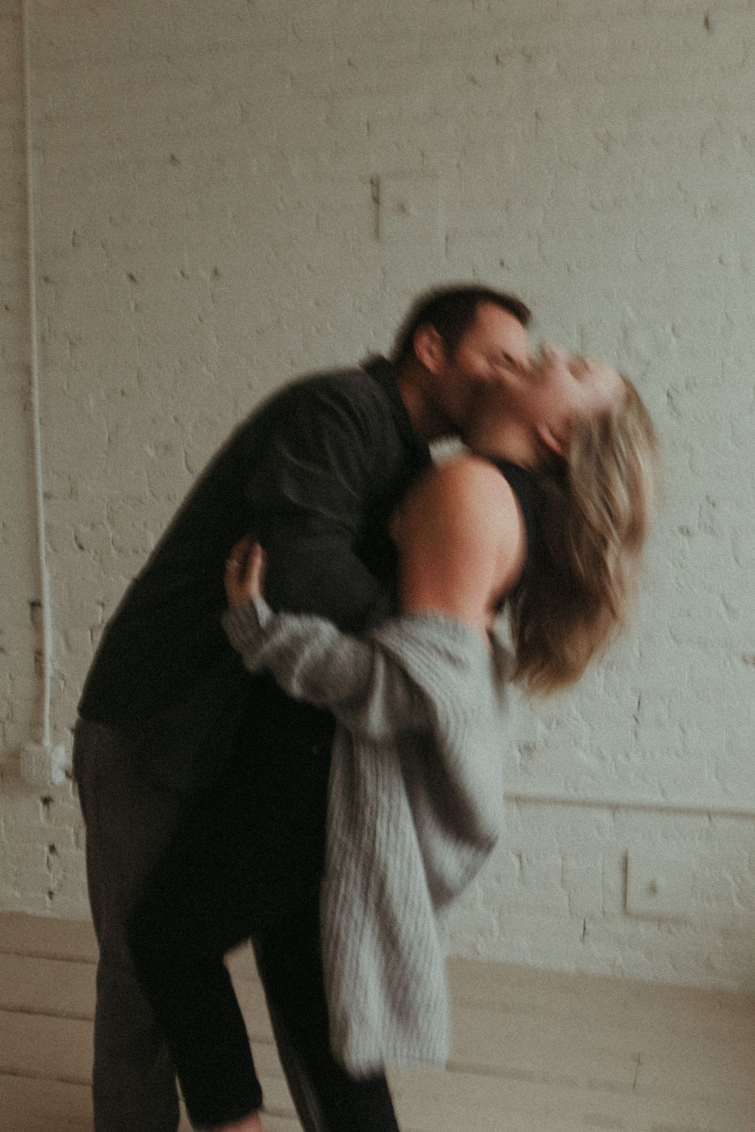 A couple shares a passionate kiss while embracing in a bright white brick room, captured in a dreamy motion blur.