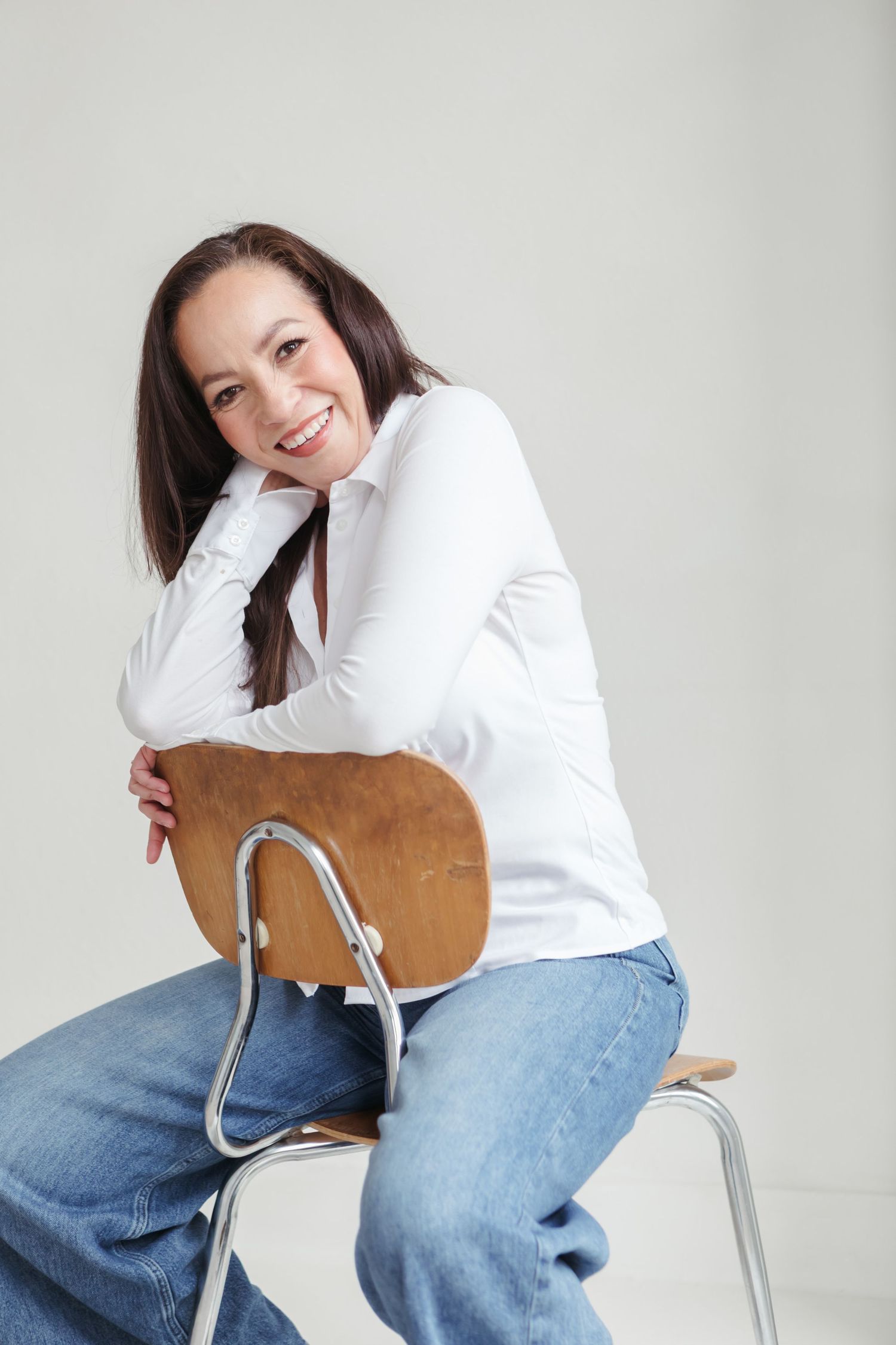 Smiling brunette woman in white top and jeans sitting backwards on a wooden chair against a light gray background.