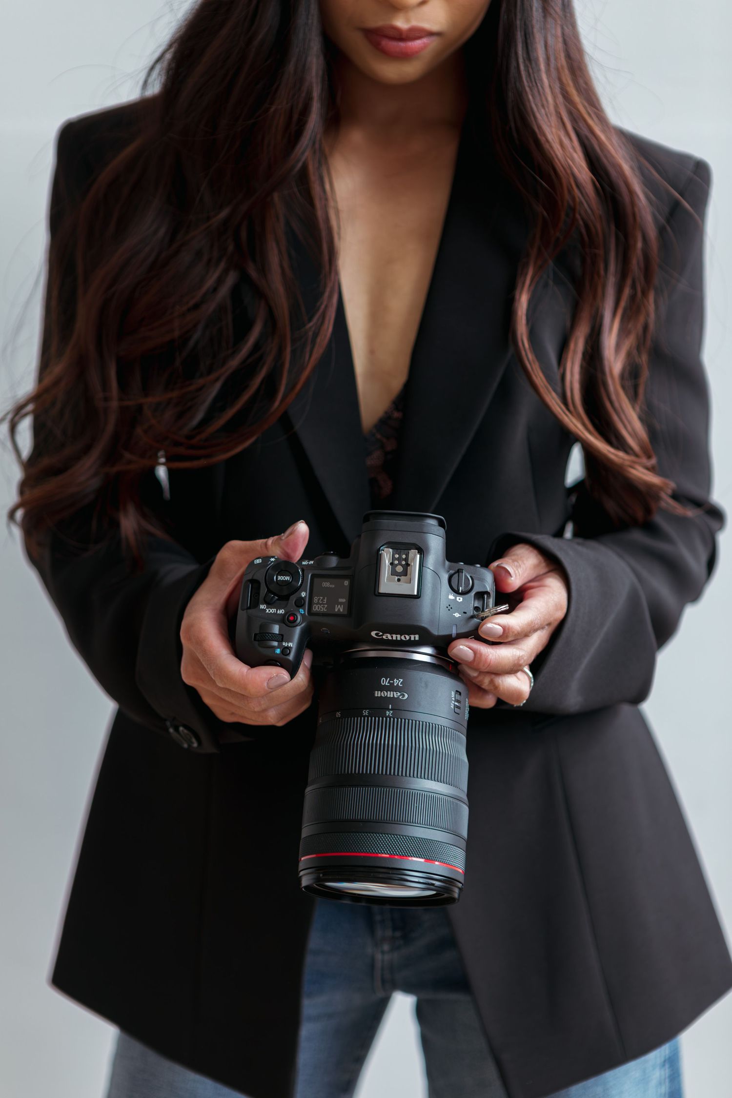 Woman with long wavy brown hair in a black blazer and jeans holding a Canon mirrorless camera with a large lens.