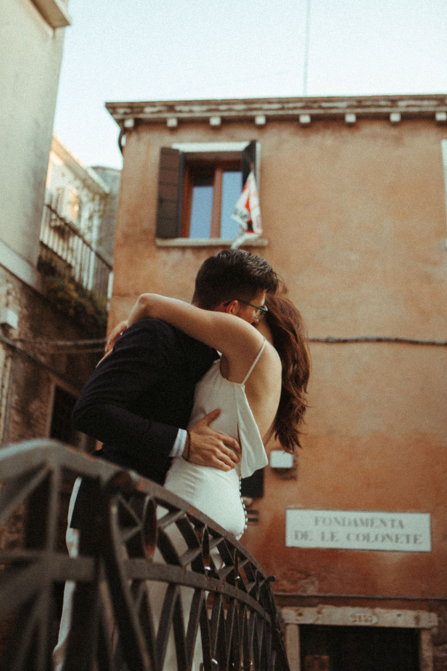 A couple embraces romantically on a bridge in Venice, Italy, with historic buildings in the background.