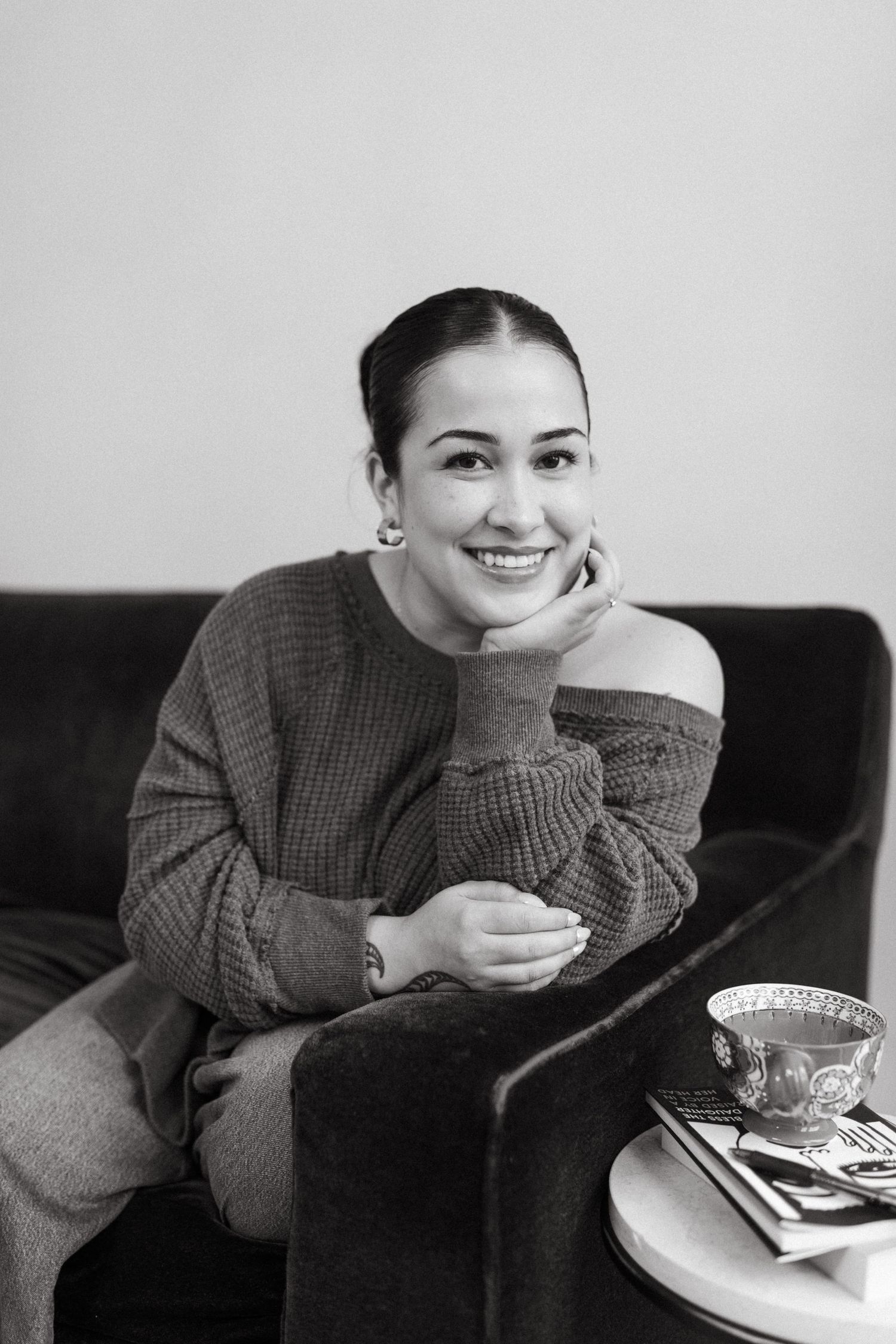 A person in a gray sweater sits on a dark couch while holding a cup and looking contemplative in black and white.