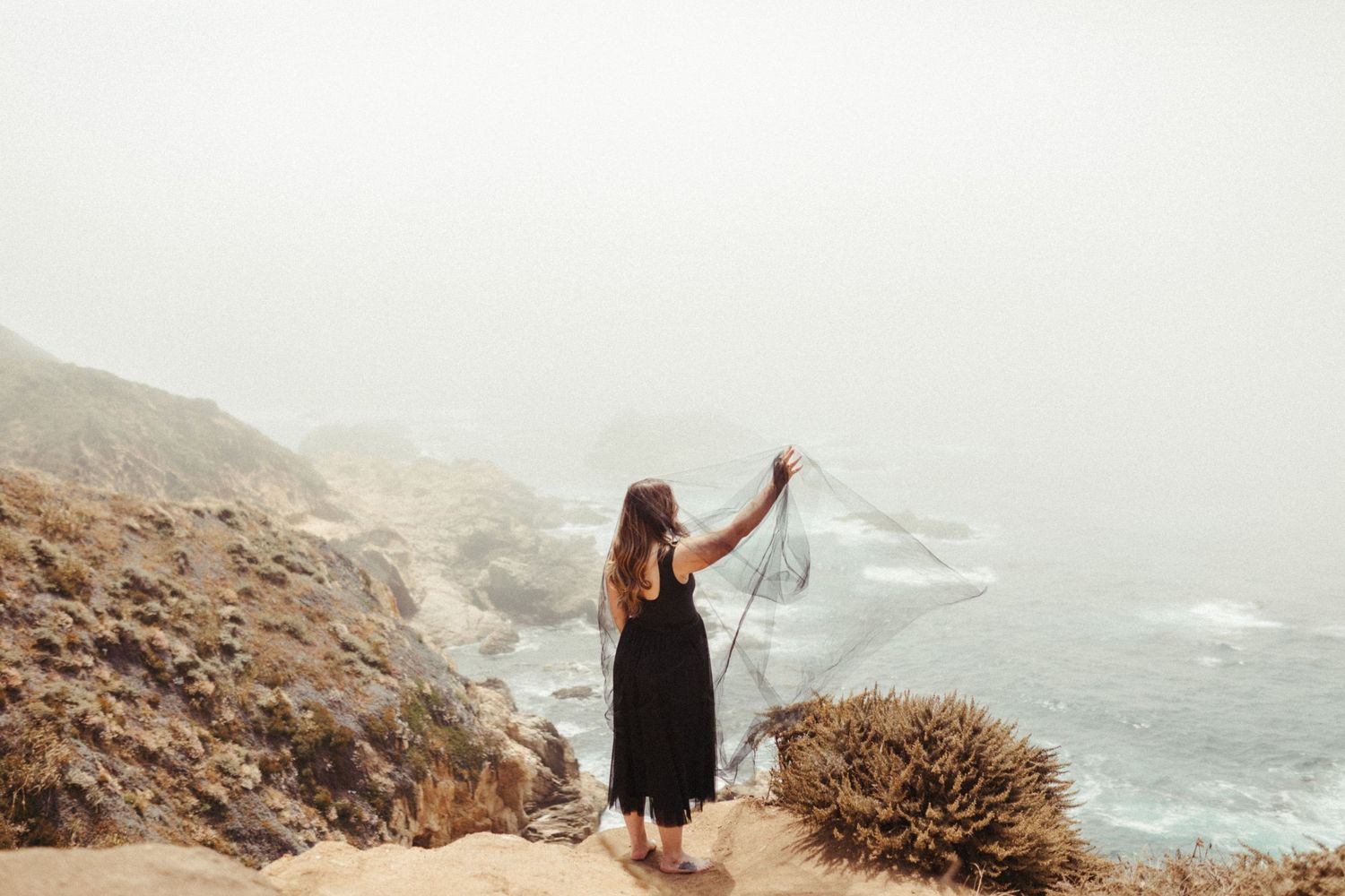 Couple stands on misty coastal cliffs overlooking the dramatic Big Sur coastline in California.
