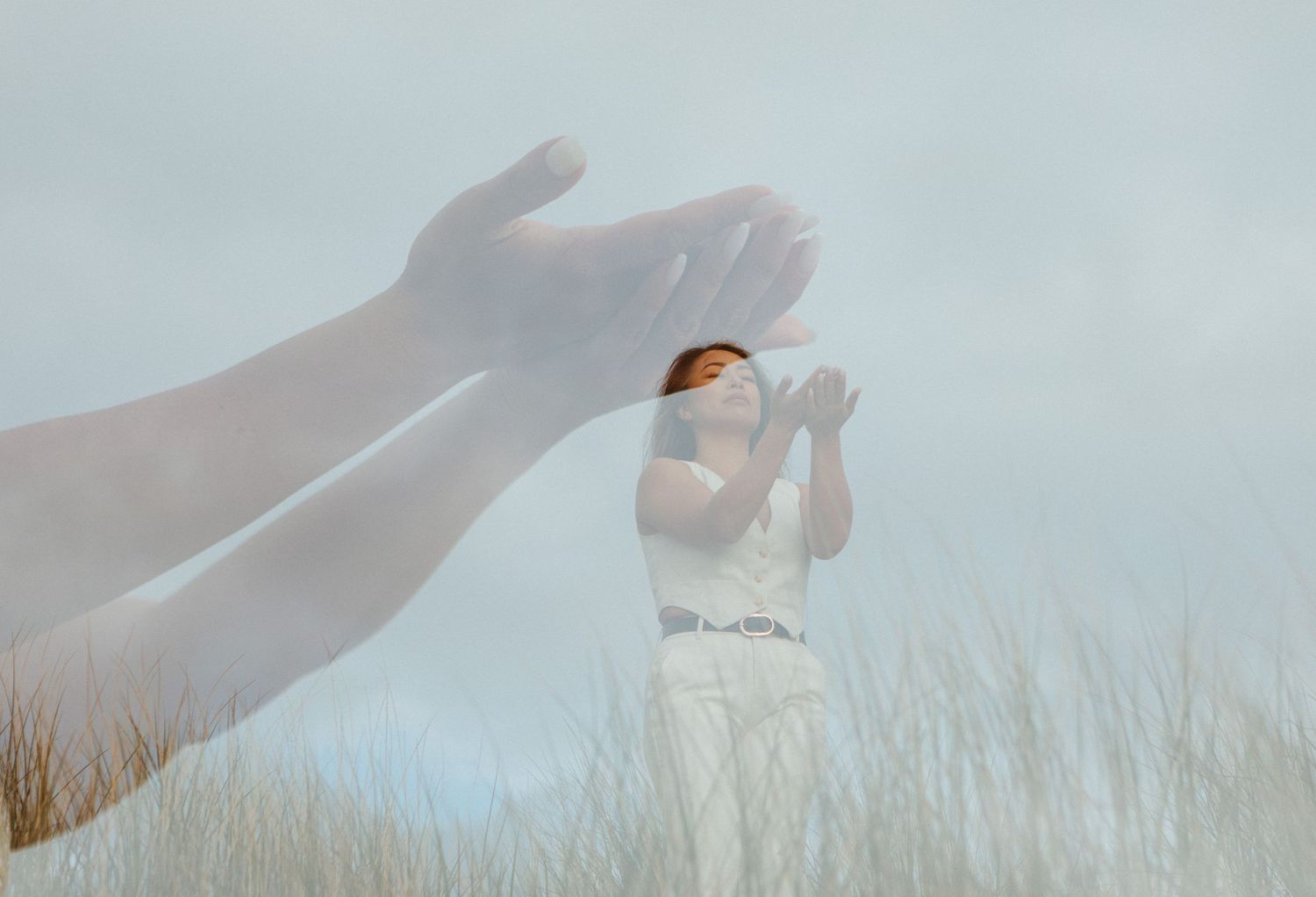 Double exposure artistic photograph showing ethereal hands reaching through tall grass against a soft cloudy sky.