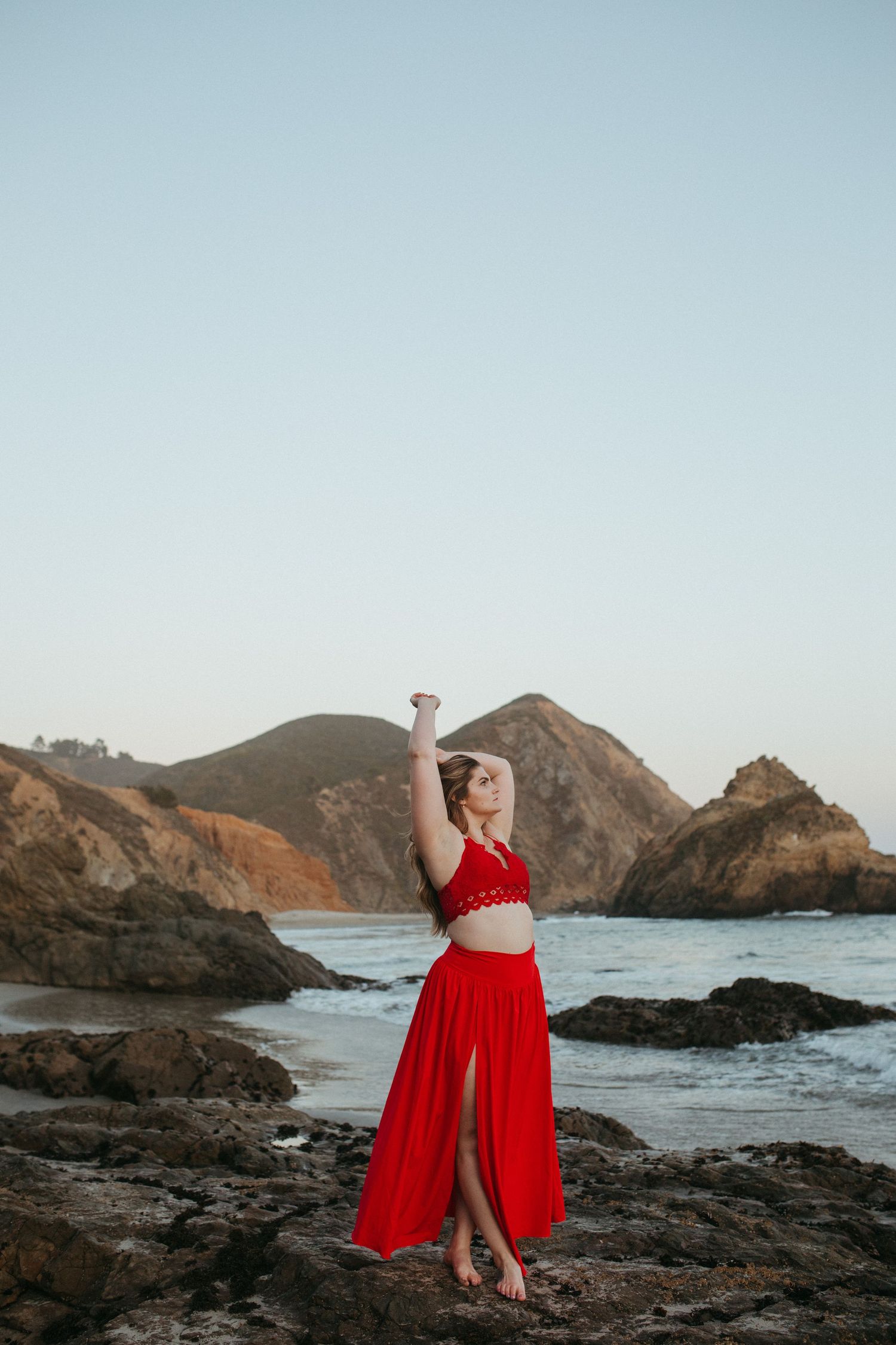 A person in a vibrant red dress poses among rocky coastal cliffs with dramatic ocean views in the background.