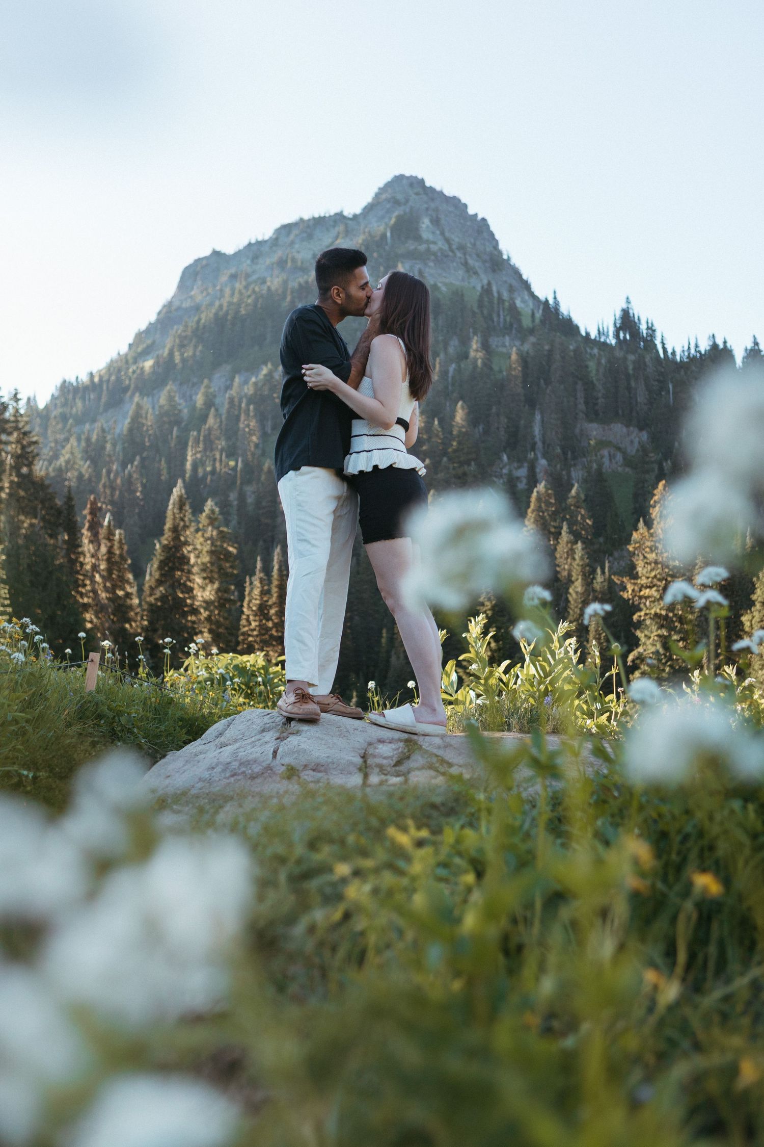 Romantic couple poses in mountain meadow with wildflowers and dramatic peaks in background.