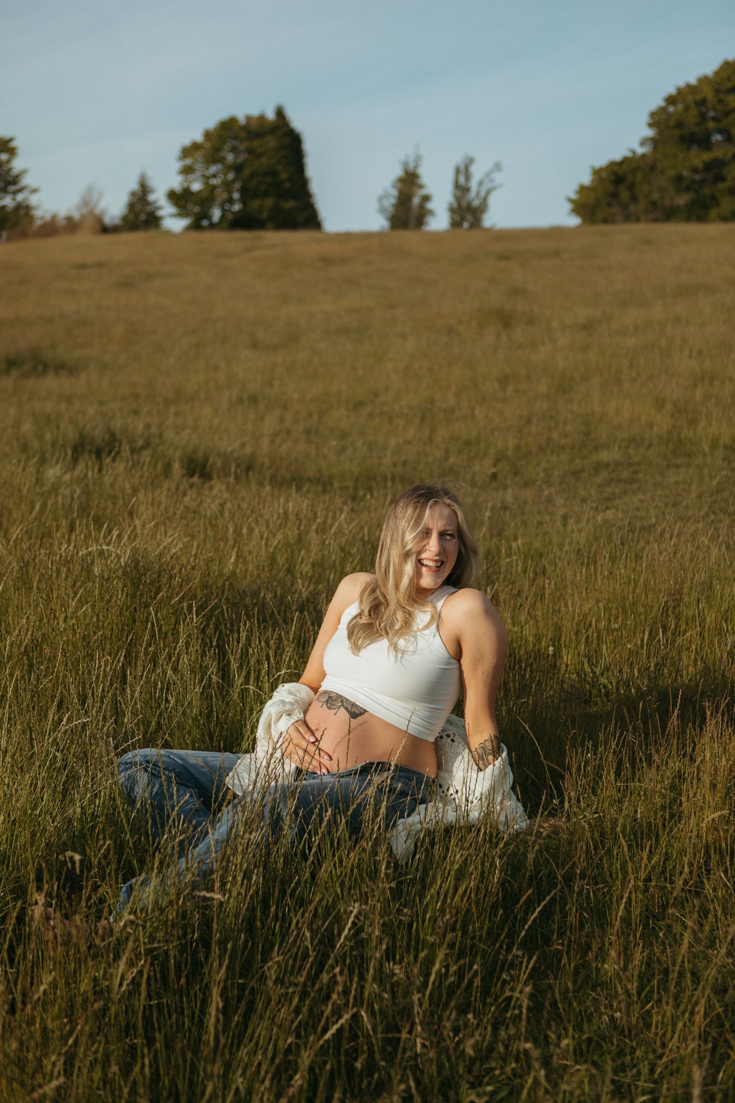 A pregnant woman in white lounges peacefully in a sunlit meadow with tall grass.