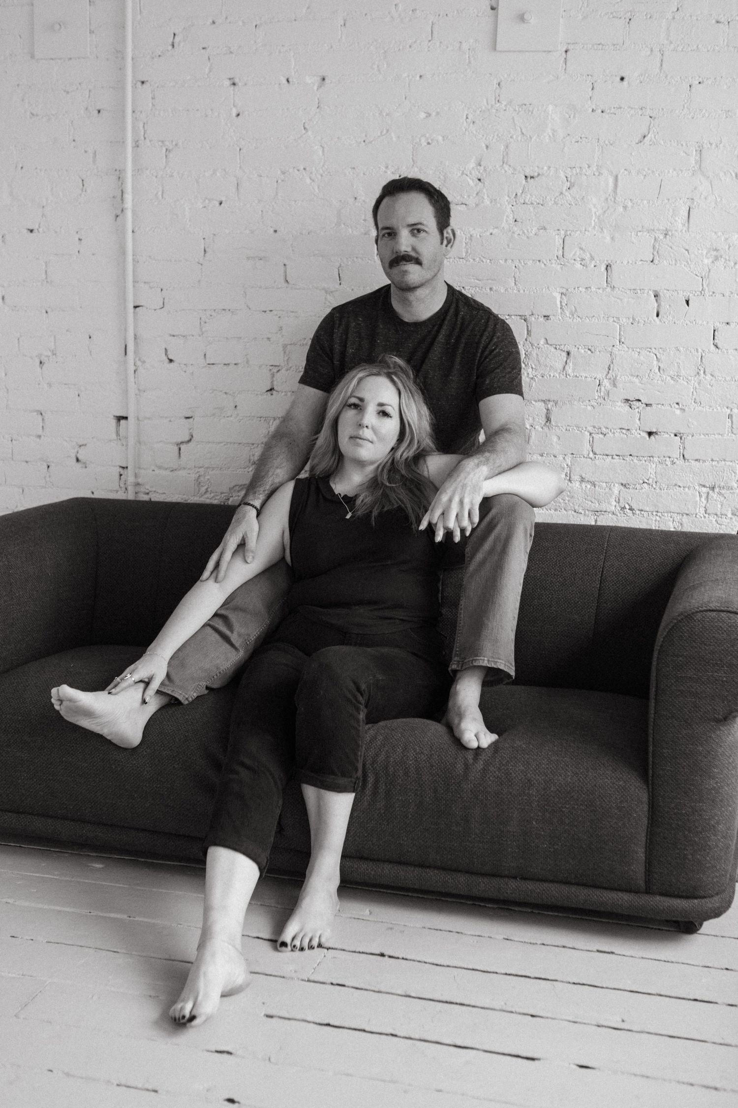 Black and white portrait of couple on gray sofa, woman seated in front with man relaxed behind her.