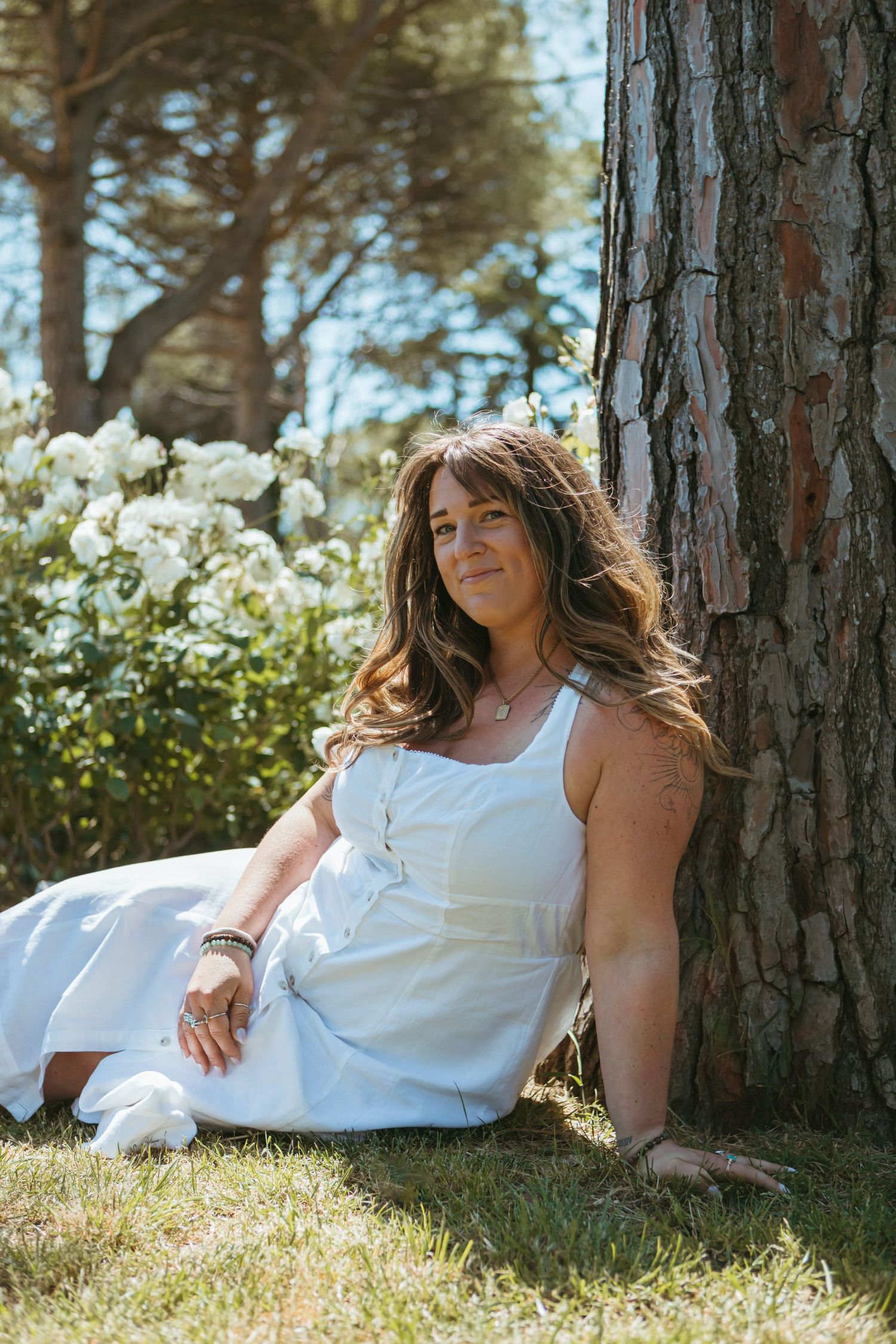 Woman in white dress sitting against tree trunk in sunny garden with white flowers and green trees.
