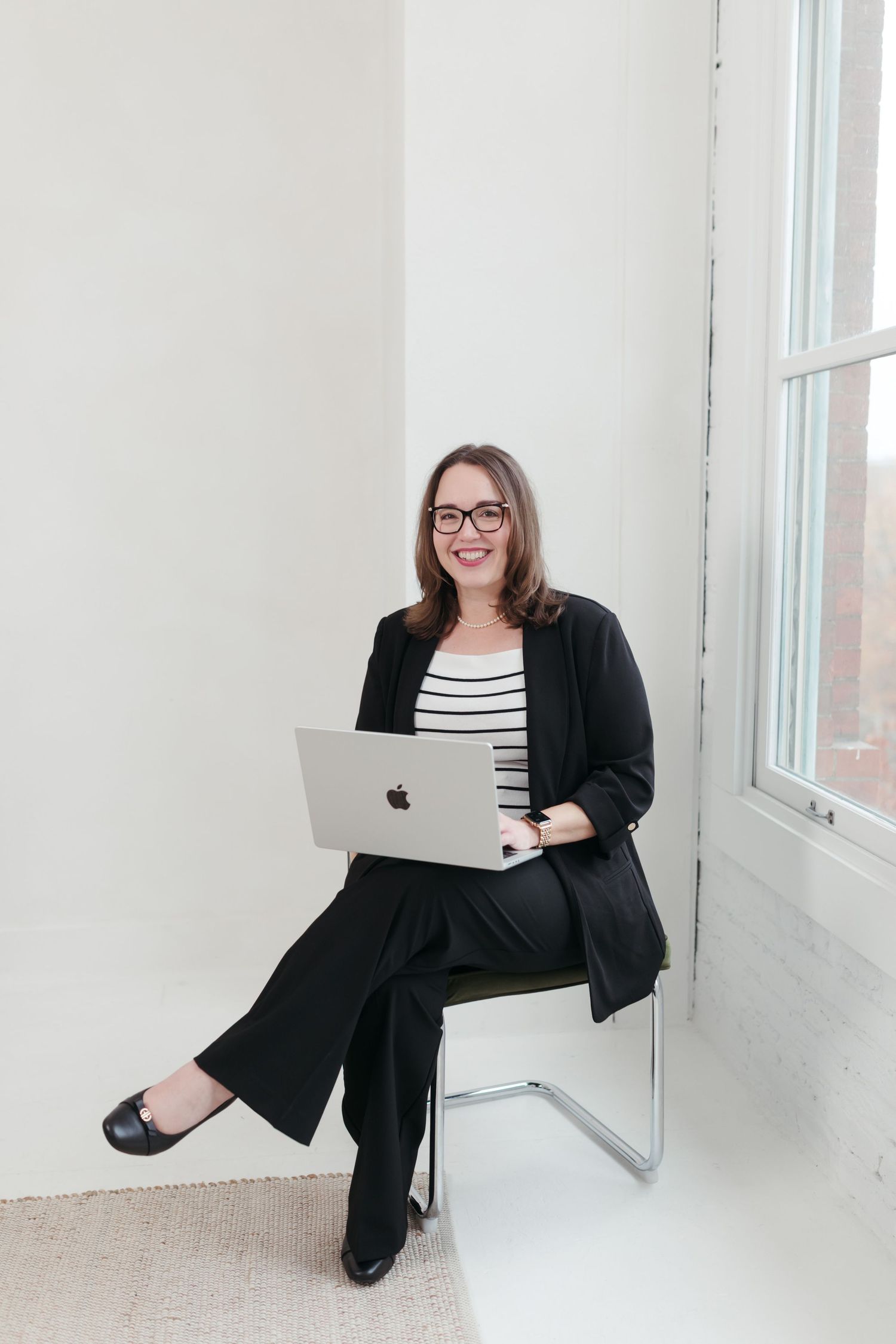 Professional wearing a black blazer and striped top poses with laptop in bright minimalist office setting.