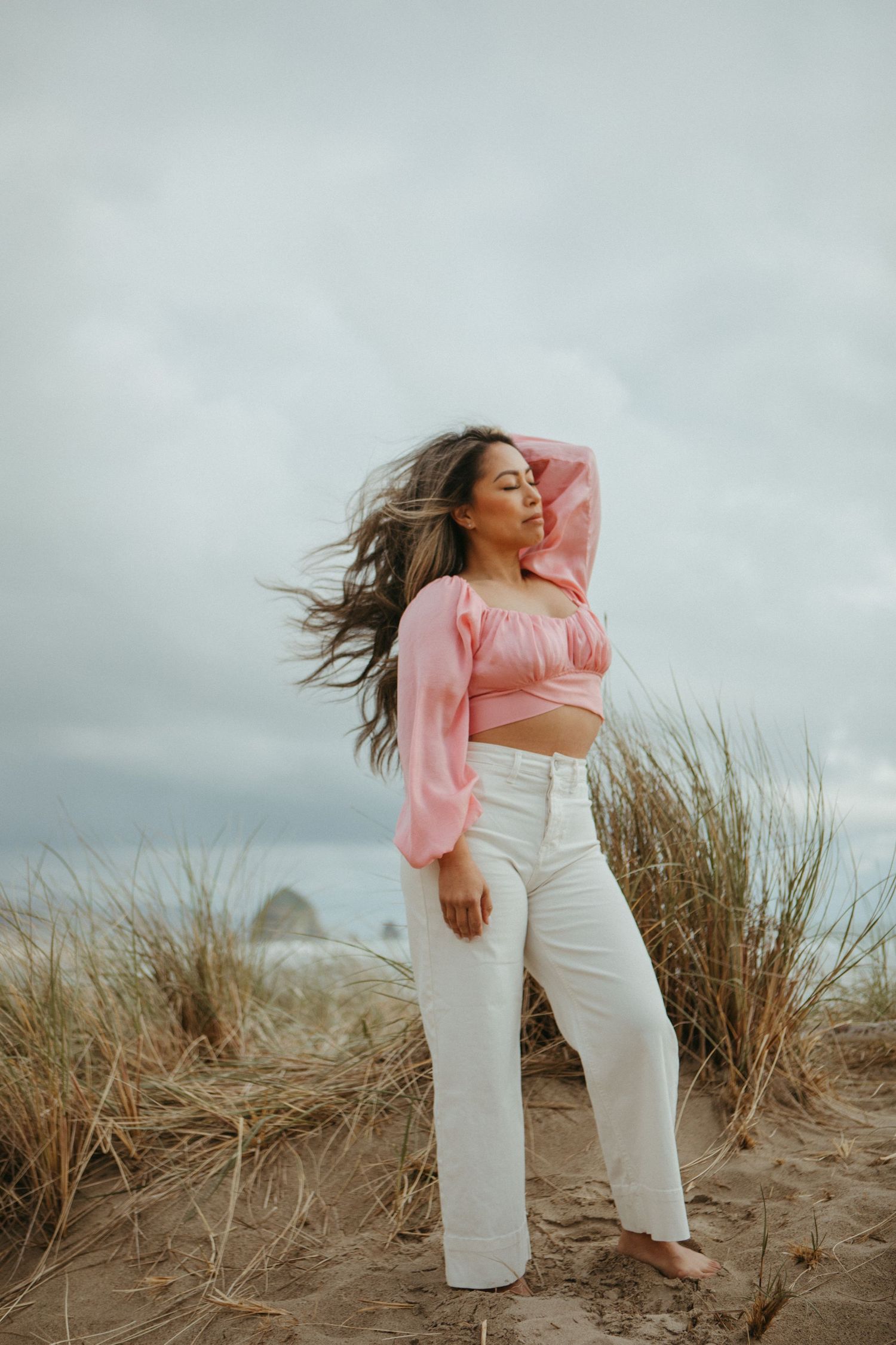 A figure in a pink sweater and white pants poses gracefully against tall beach grass and cloudy skies.