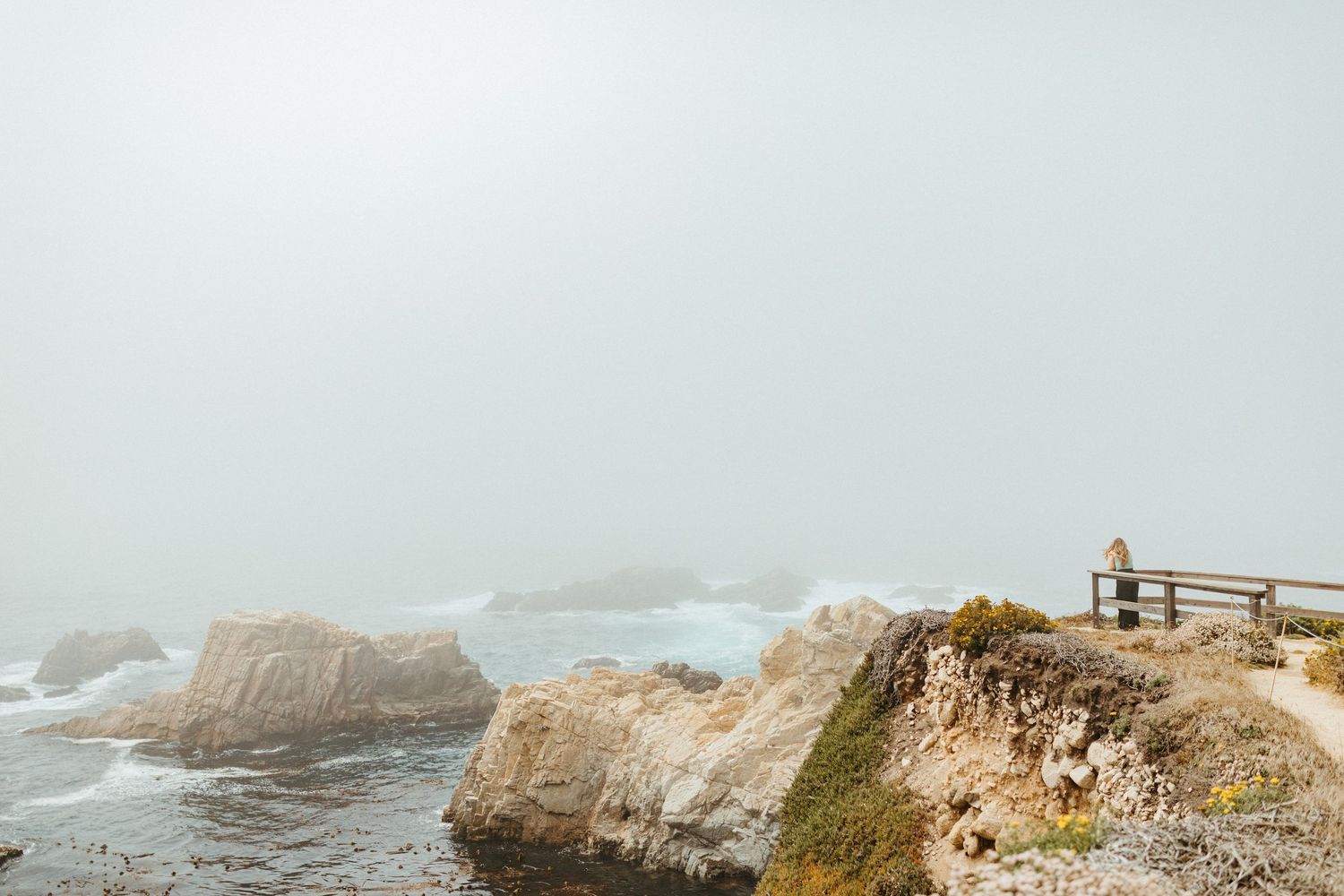 Scenic coastal view of rocky cliffs overlooking a misty ocean with people walking along a fenced path.