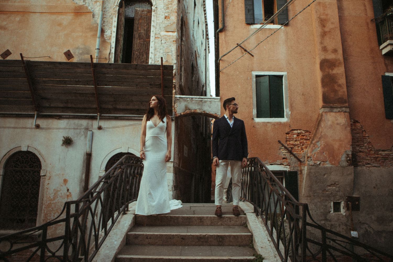 Bride and groom stand on stone bridge steps in Venice, Italy, surrounded by rustic aged buildings.