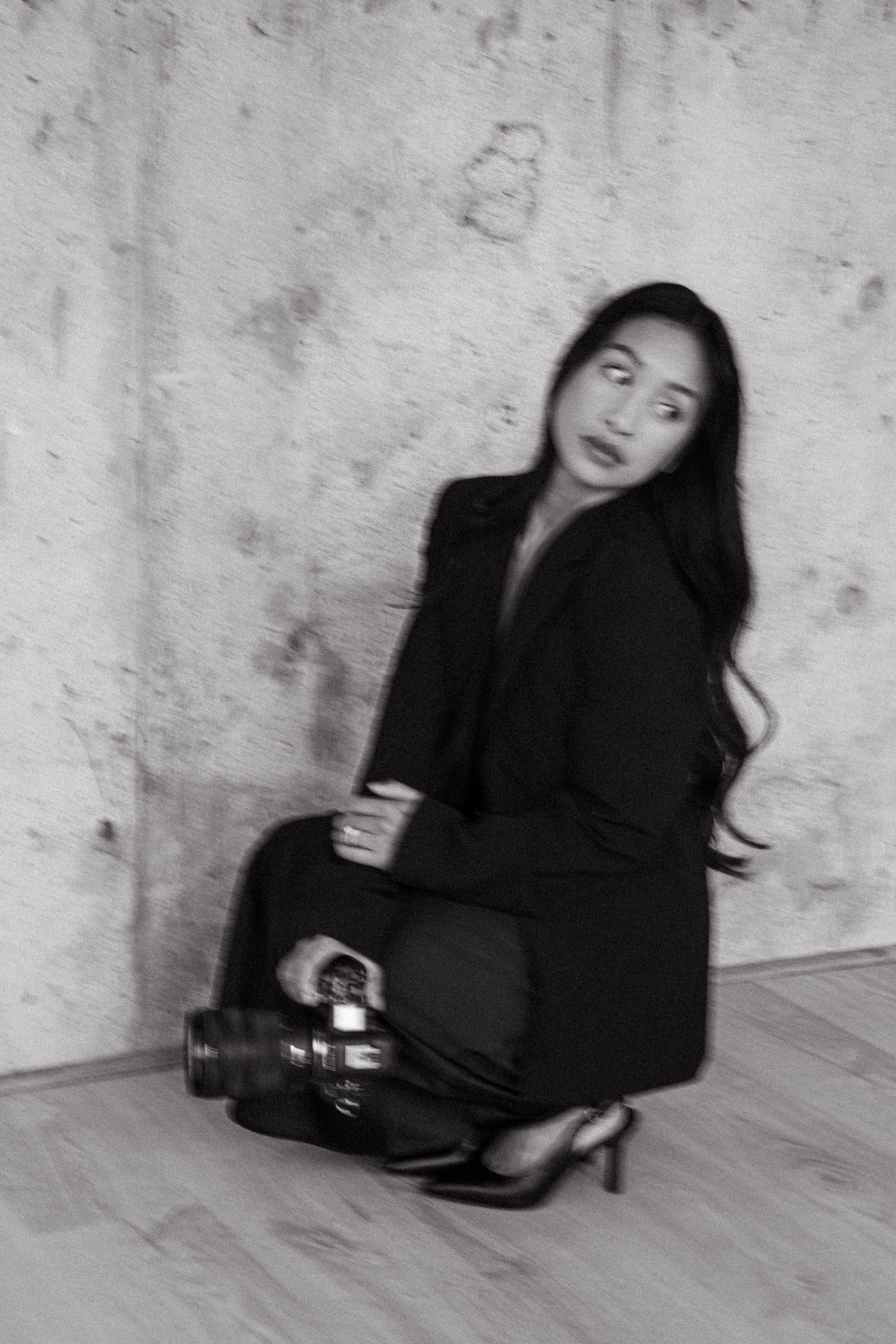 Black and white photo of a woman in a blazer and heels crouching against a concrete wall, holding a camera and glancing over her shoulder.