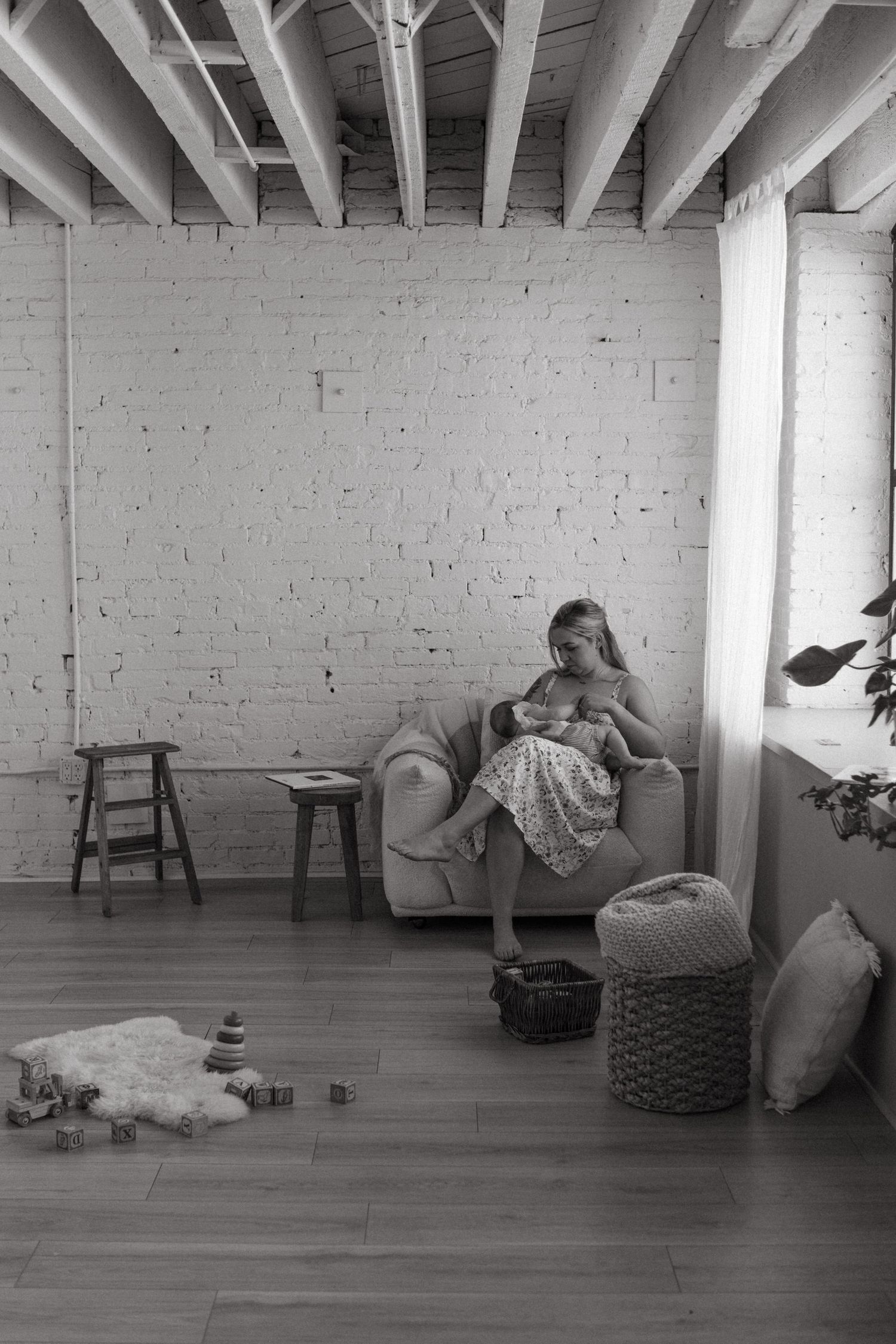 Black and white photo of a mother breastfeeding her baby in an armchair in a rustic loft-style room.