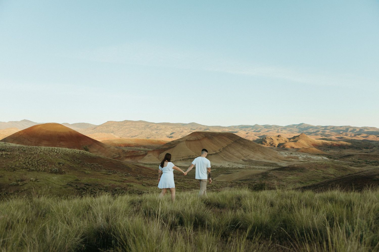 Couple holding hands in a vast golden grassland with rolling desert hills under a clear blue sky.