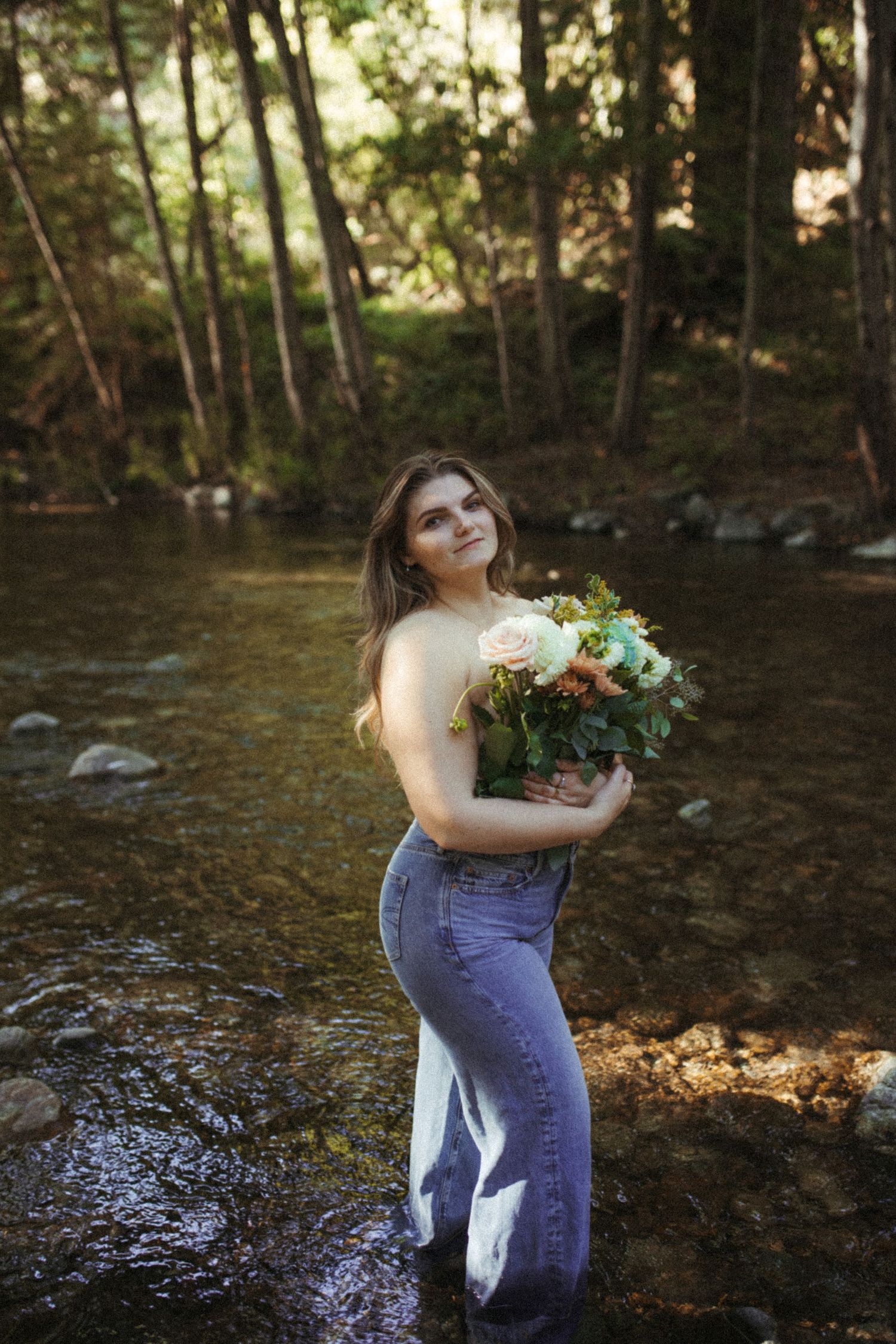 A person holding a vibrant bouquet poses by a forest stream wearing jeans and a sleeveless top.