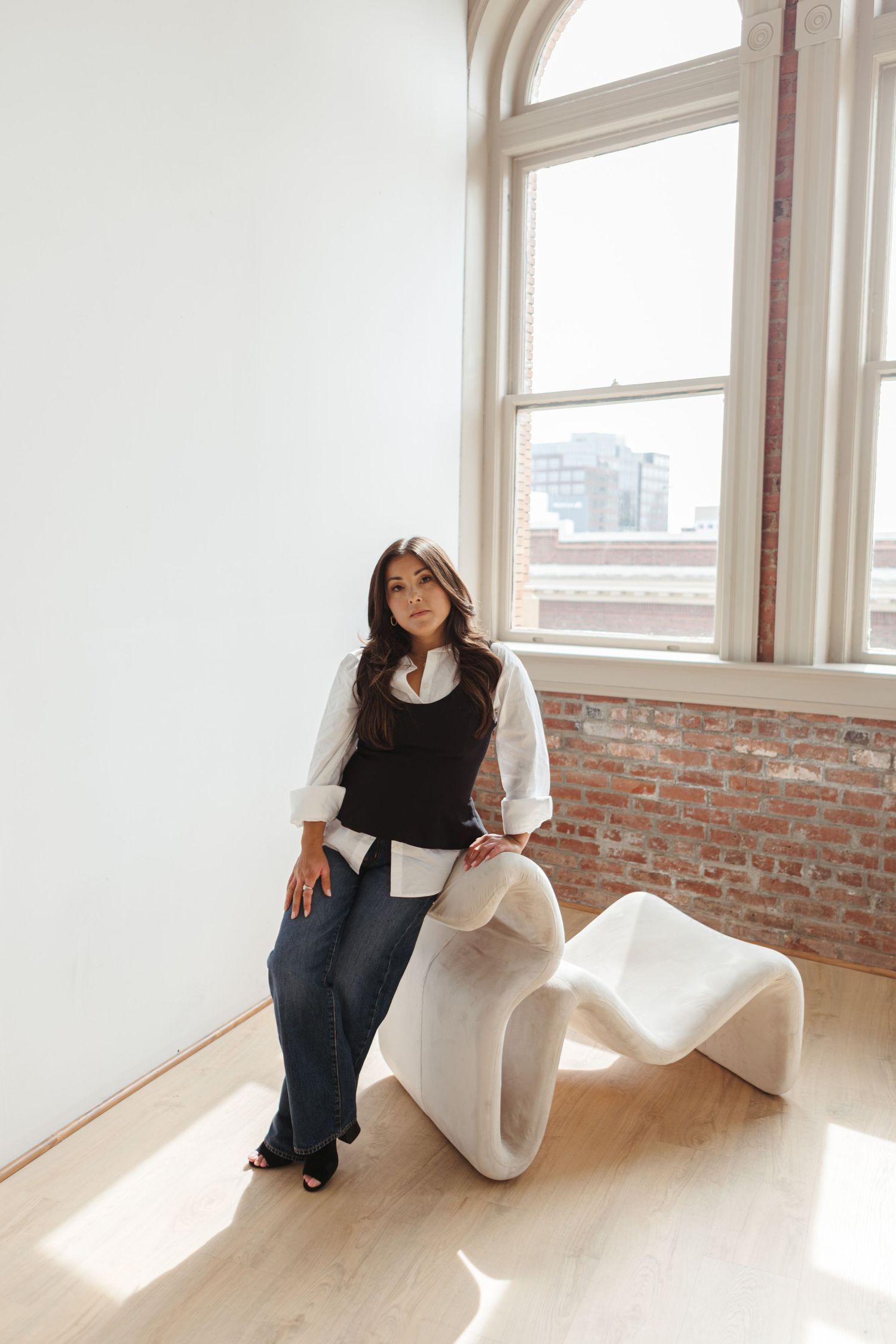 Woman in a black vest, white shirt and wide-leg jeans sits on a sculptural white chair in a bright loft with brick walls and large windows.