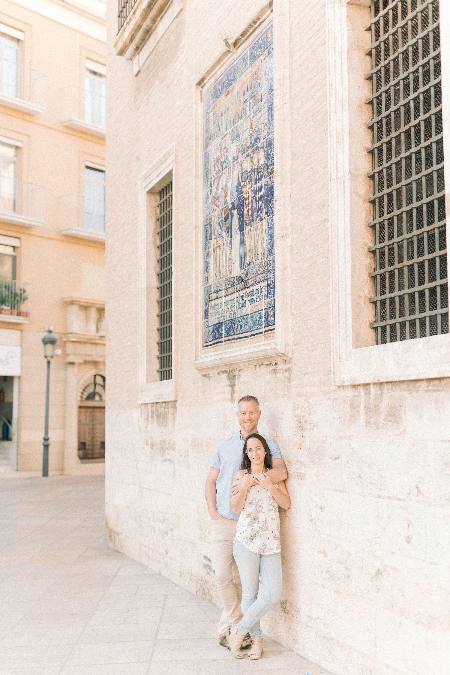 A couple poses against a historic stone building with decorative blue tile artwork in a Mediterranean setting.