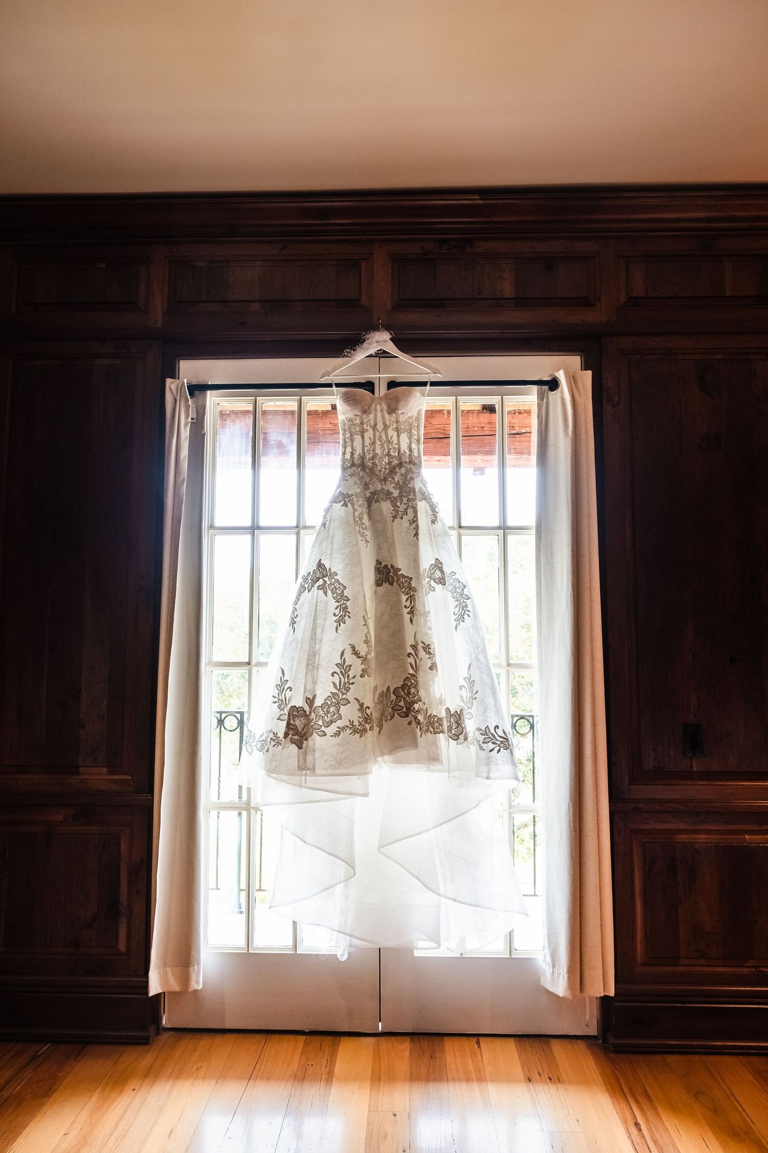 A wedding dress hangs illuminated in front of a bright window creating a silhouette against dark wood paneled walls. Monterey California.