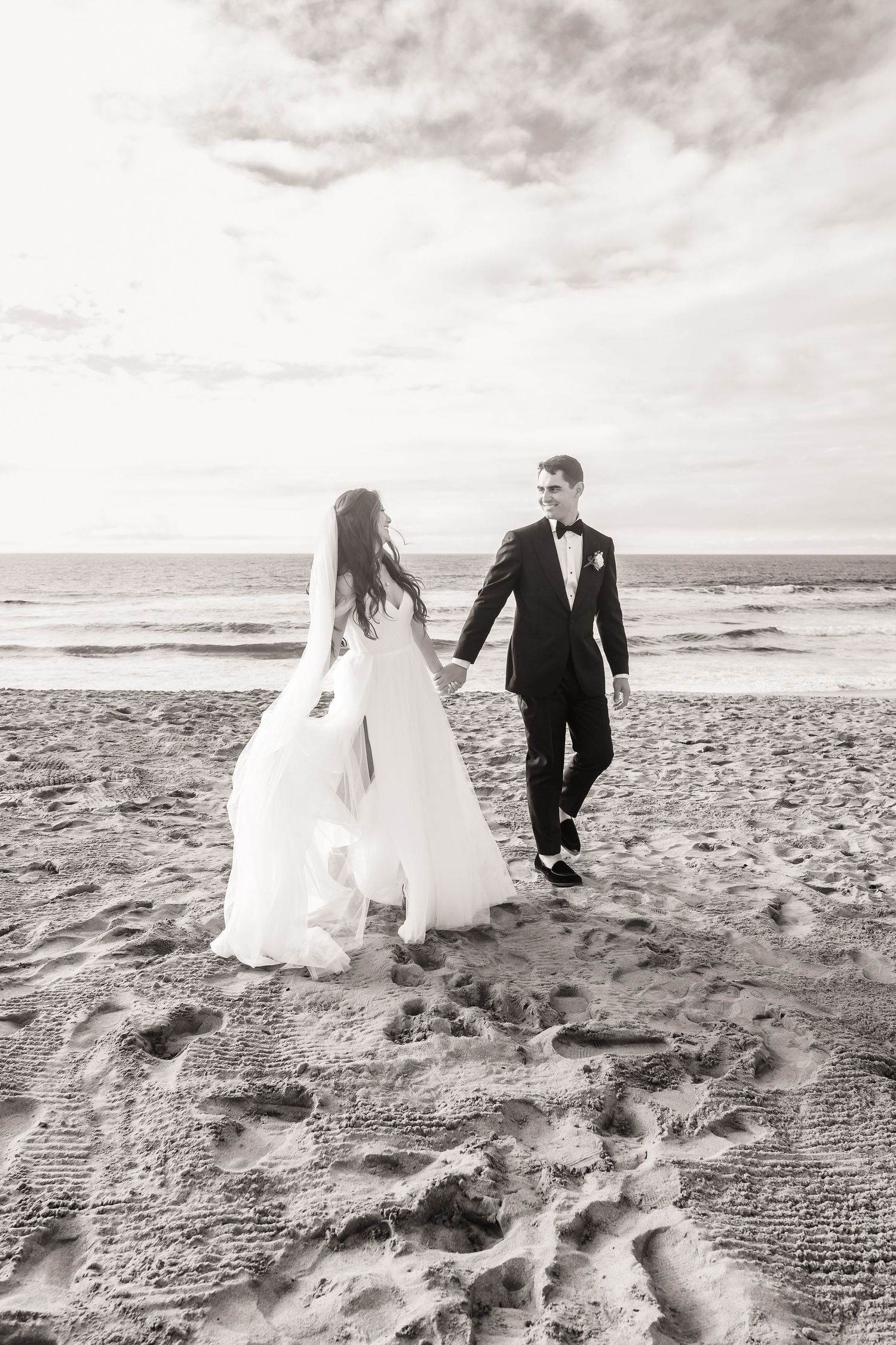 A bride and groom walk along a rocky beach shoreline in black and white photography on their wedding day in Monterey at The Sanctuary Beach