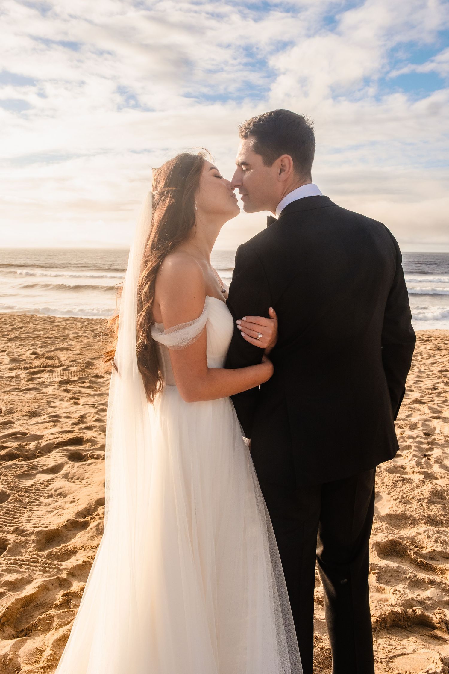 A romantic beach wedding at The Sanctuary Beach shows a couple embracing on sandy shores with ocean waves and cloudy skies in the background
