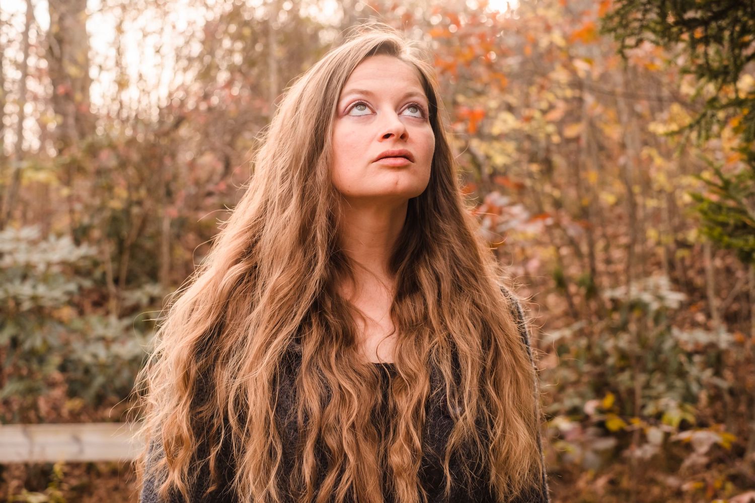 Long wavy hair flows in autumn forest setting during golden hour with warm fall colors in background.