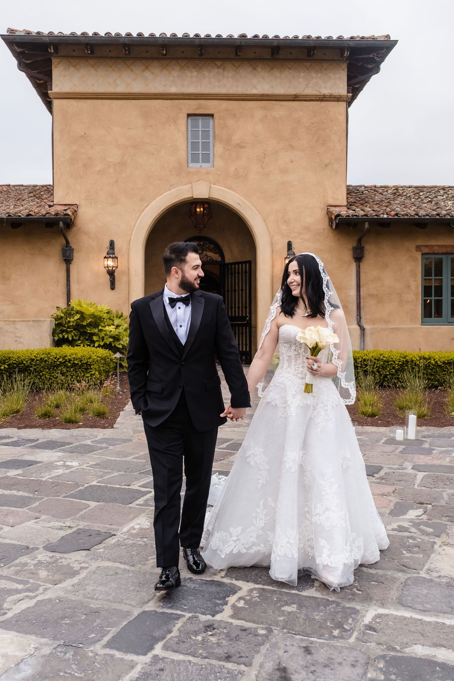 A bride and groom walk together along a stone courtyard at a Spanish colonial style mission building. A Monterey California wedding venue.
