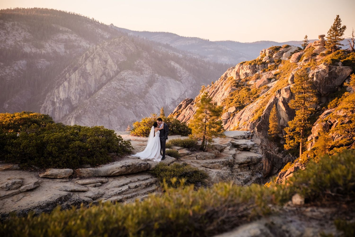 A couple stands on a rocky overlook at sunset in Yosemite National Park with majestic mountain views in the background.