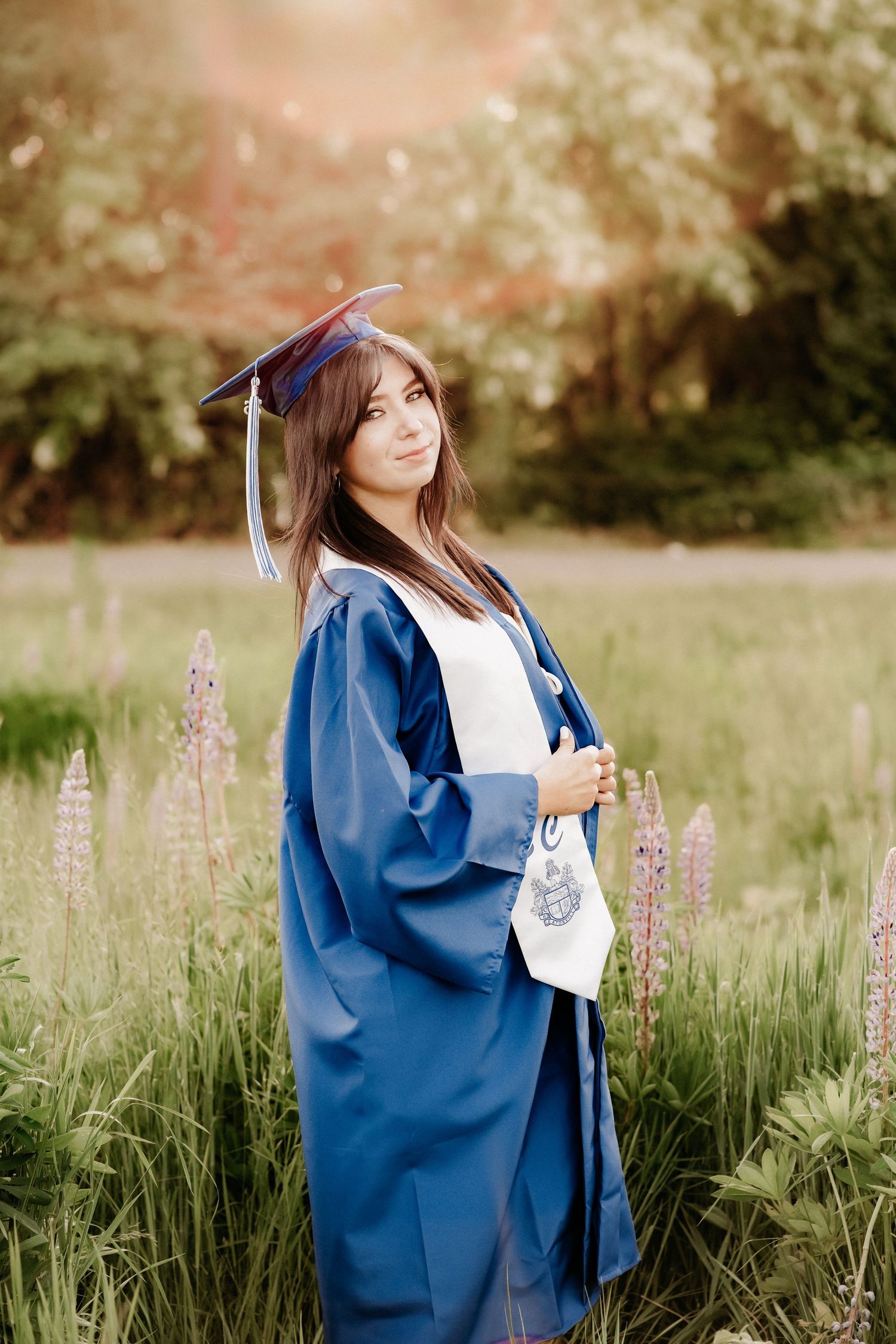 Young woman in blue graduation cap and gown posing outdoors among wildflowers in soft sunlight during senior photo session in Puyallup, Wa.