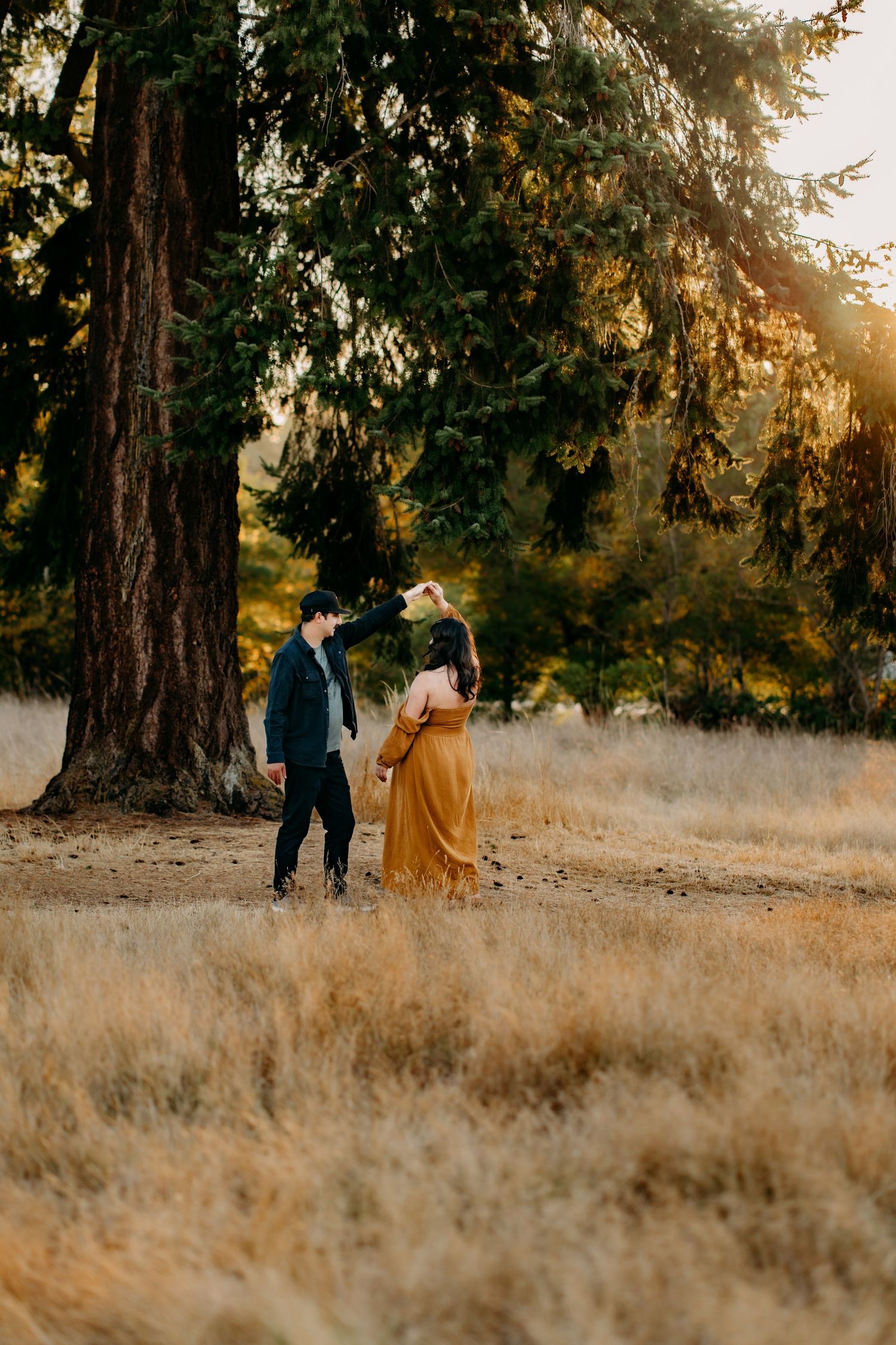 Couple dances in a golden field at sunset with tall evergreen trees creating a dreamy autumn atmosphere for maternity photos in Tacoma.