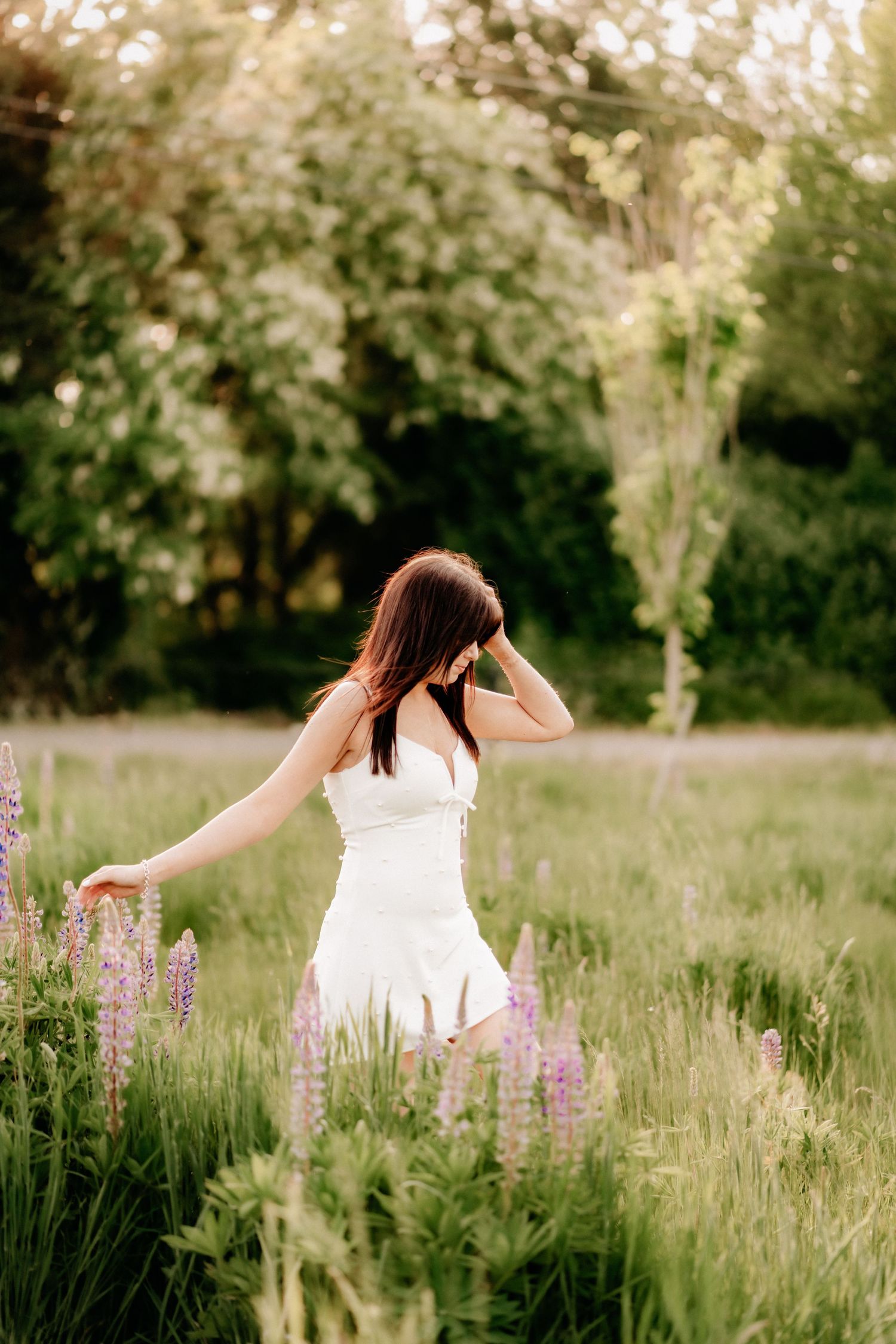 Woman in white dress walking through a field of purple lupine flowers at golden hour for high school senor photos in Puyallup, WA.