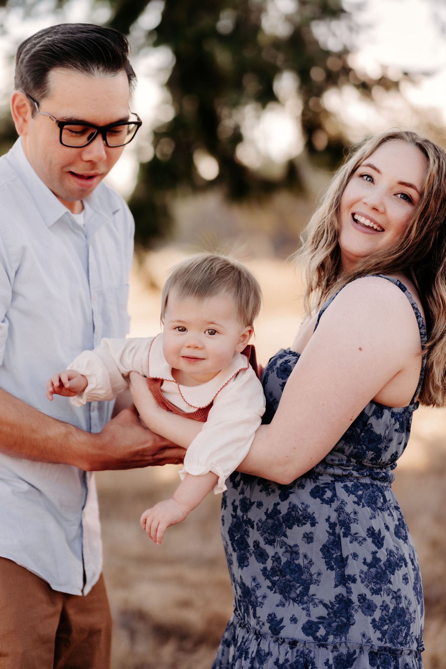 Happy family portrait of parents holding smiling baby outdoors in warm golden light during family photos in Tacoma, WA.