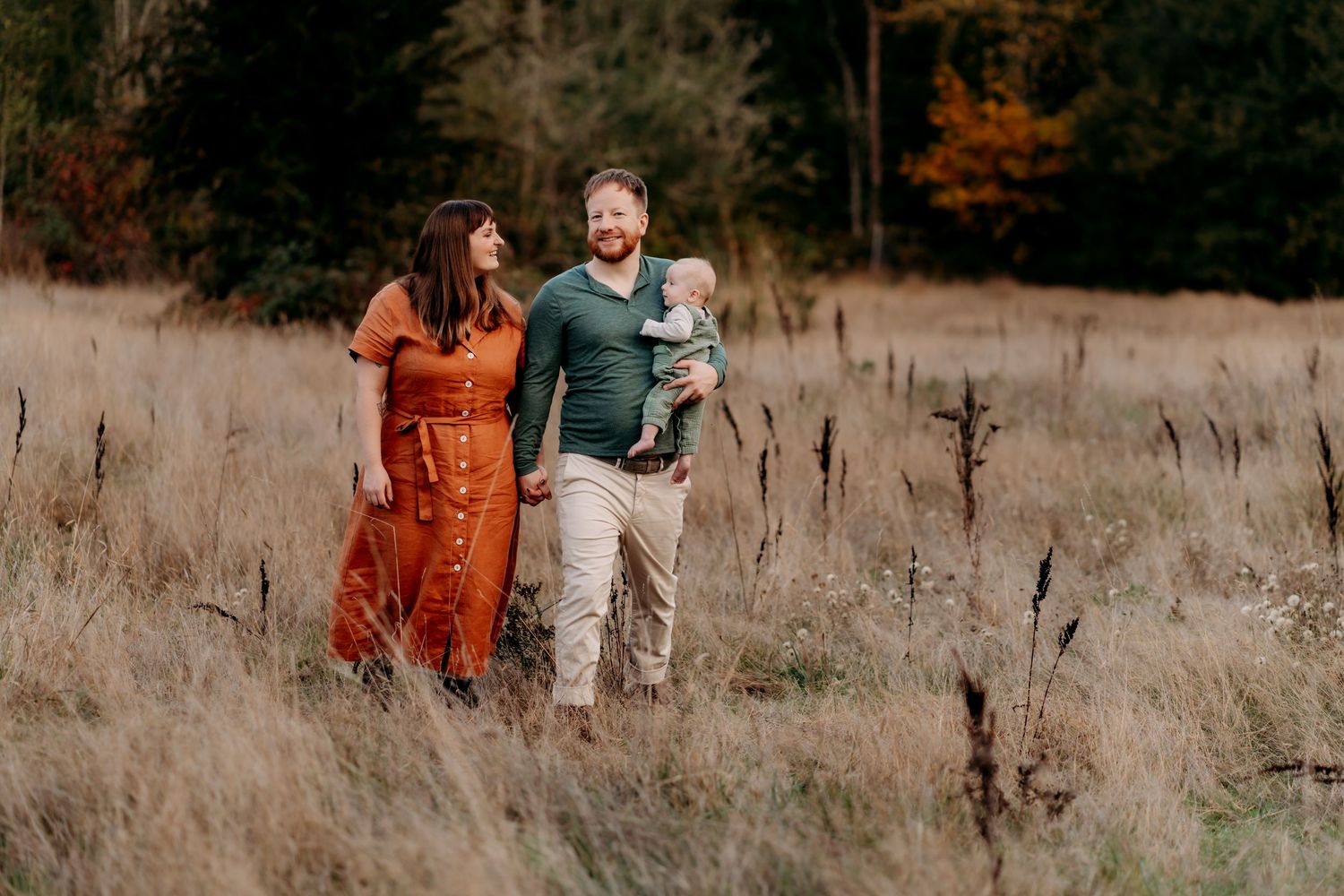 A happy family walks through a golden meadow in autumn, parents holding hands with dad carrying their baby during fall family photos.